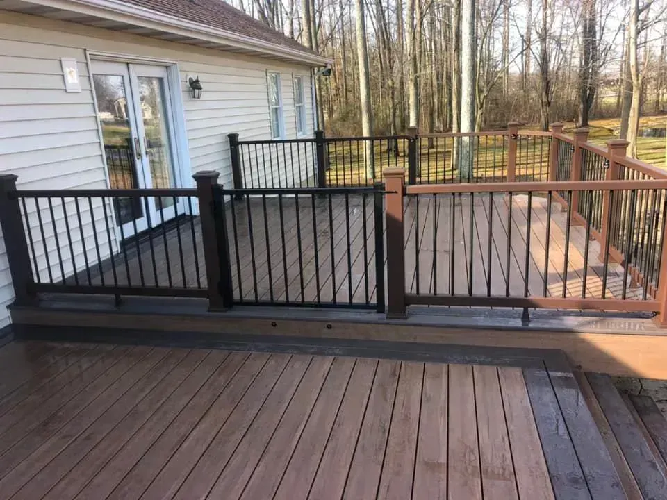 Wooden deck with dark and light brown railings and flooring next to a white house with glass doors.