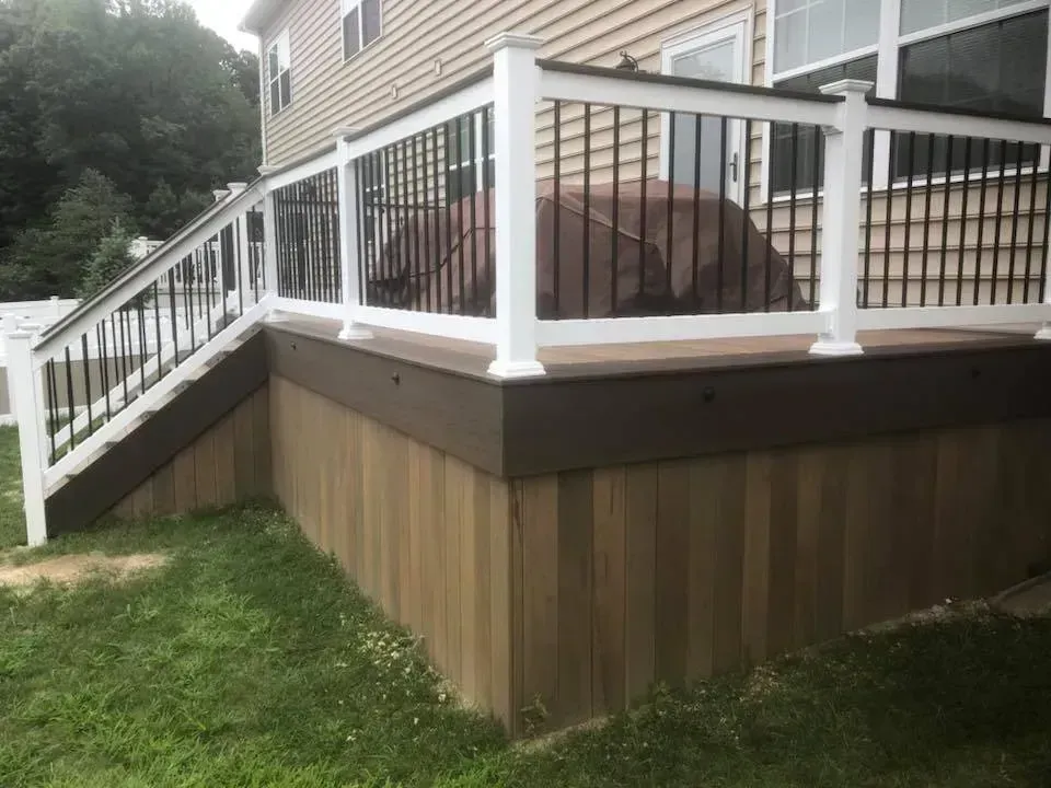 Wooden deck with white railings, black balusters, brown skirting, and stairs on green grass.