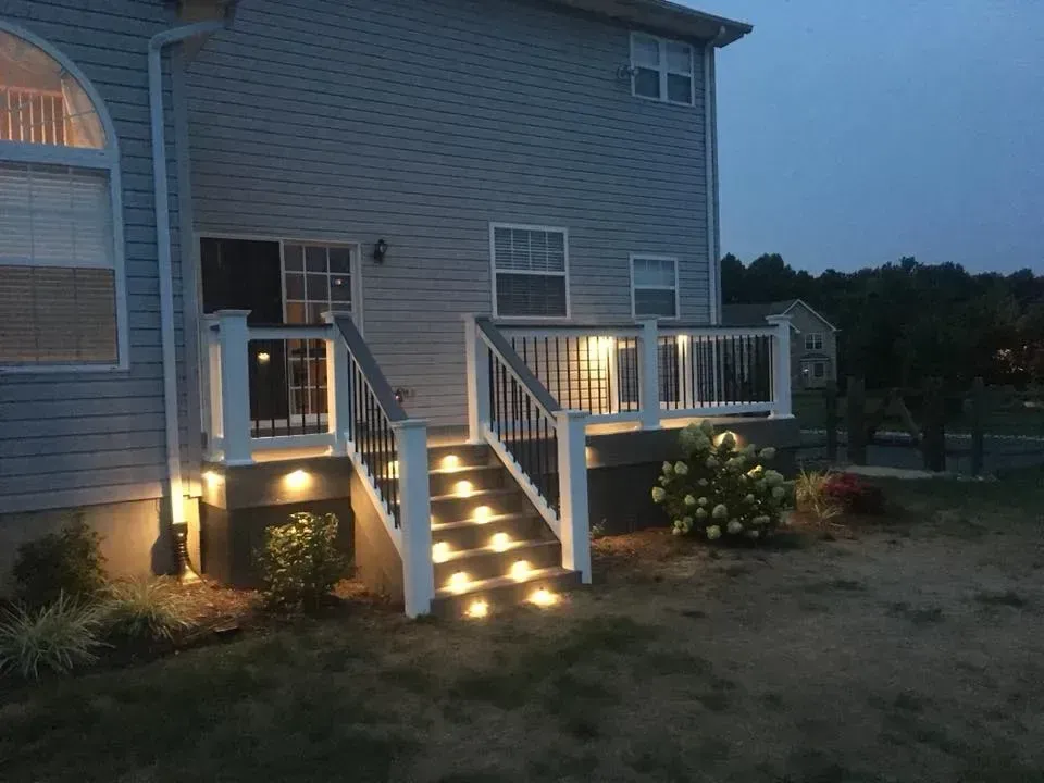 Lit deck and stairs at dusk. Exterior of a house with white railing, built-in lights, and landscaping.