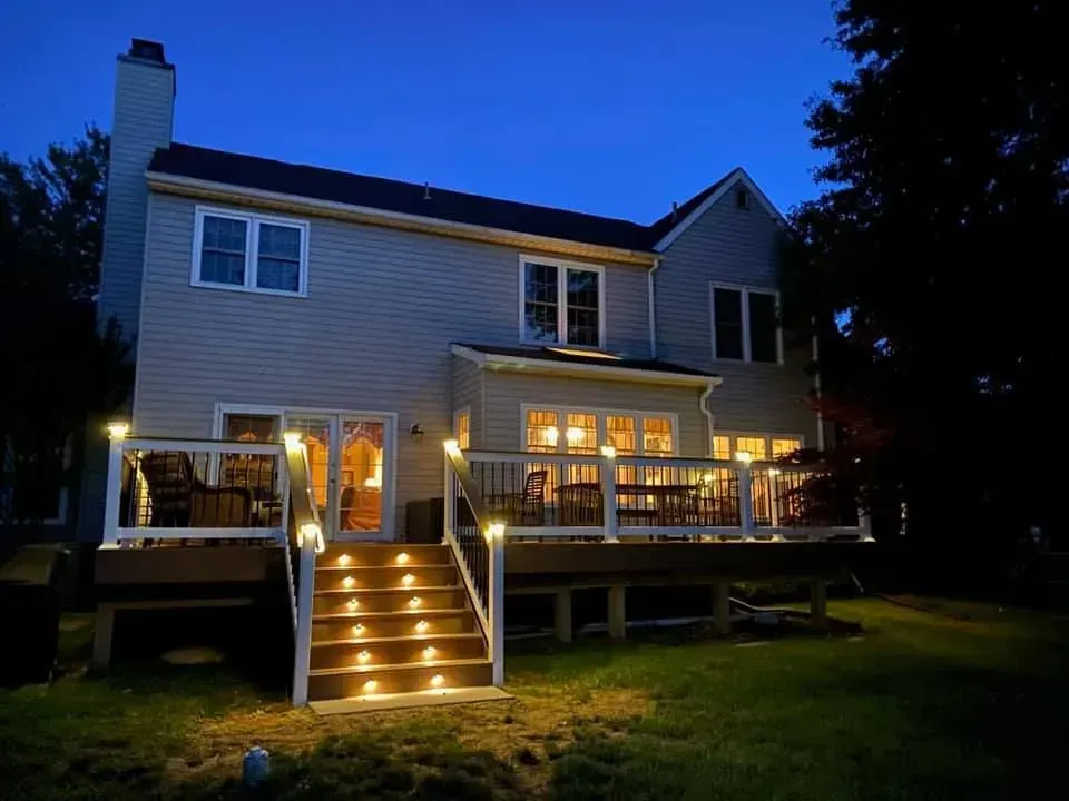 Back of a two-story house with a lit deck and stairs, at dusk. Deck and stair lights illuminate the house.