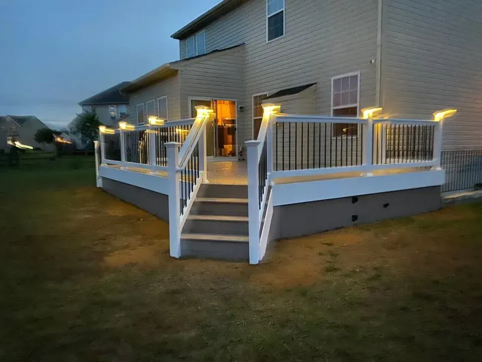 Lit deck with white railings, steps, and gray decking, attached to a tan house at dusk.