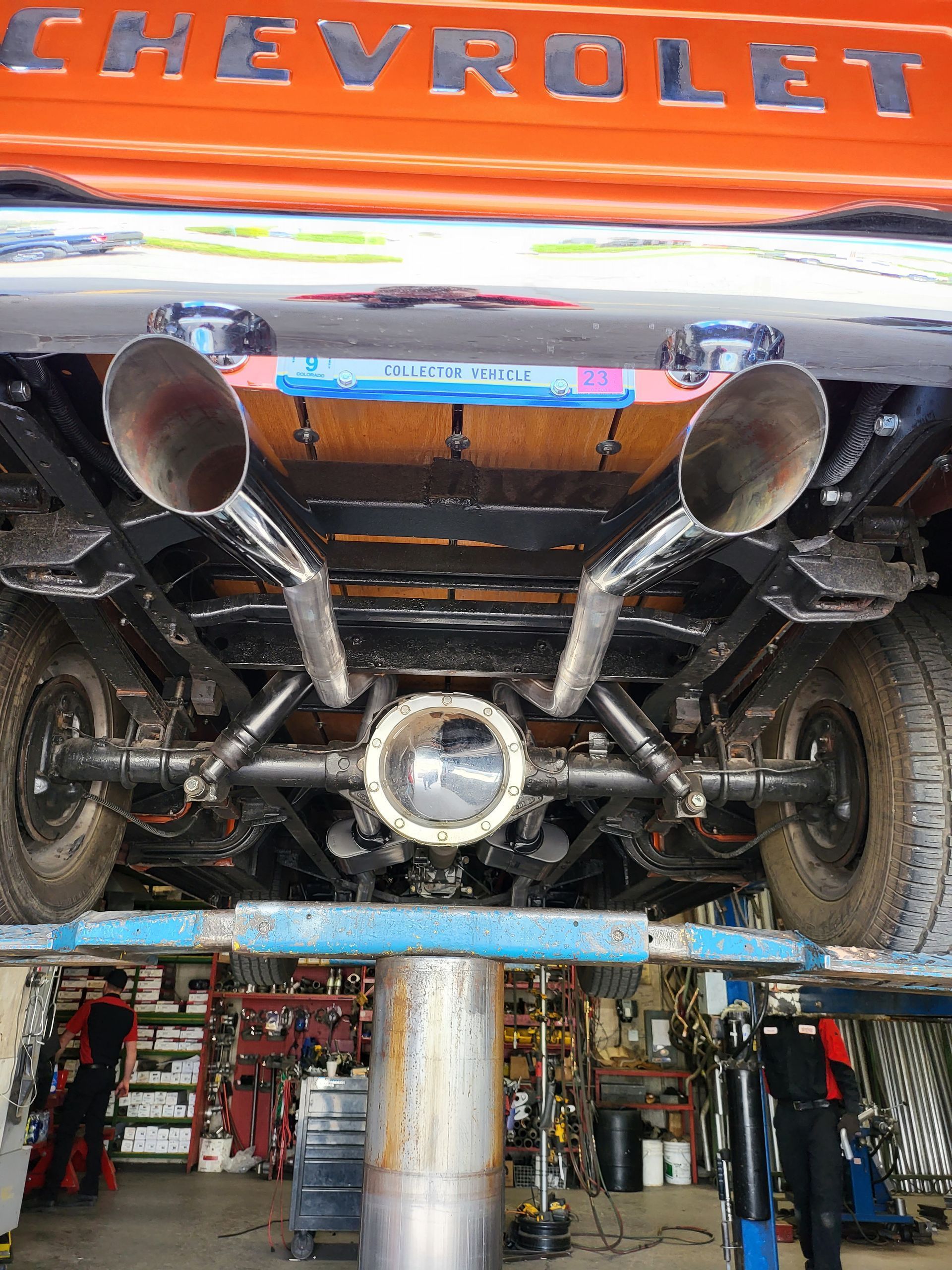 Underside view of an orange Chevrolet truck on a lift, showing custom exhaust, axles, and undercarriage details.