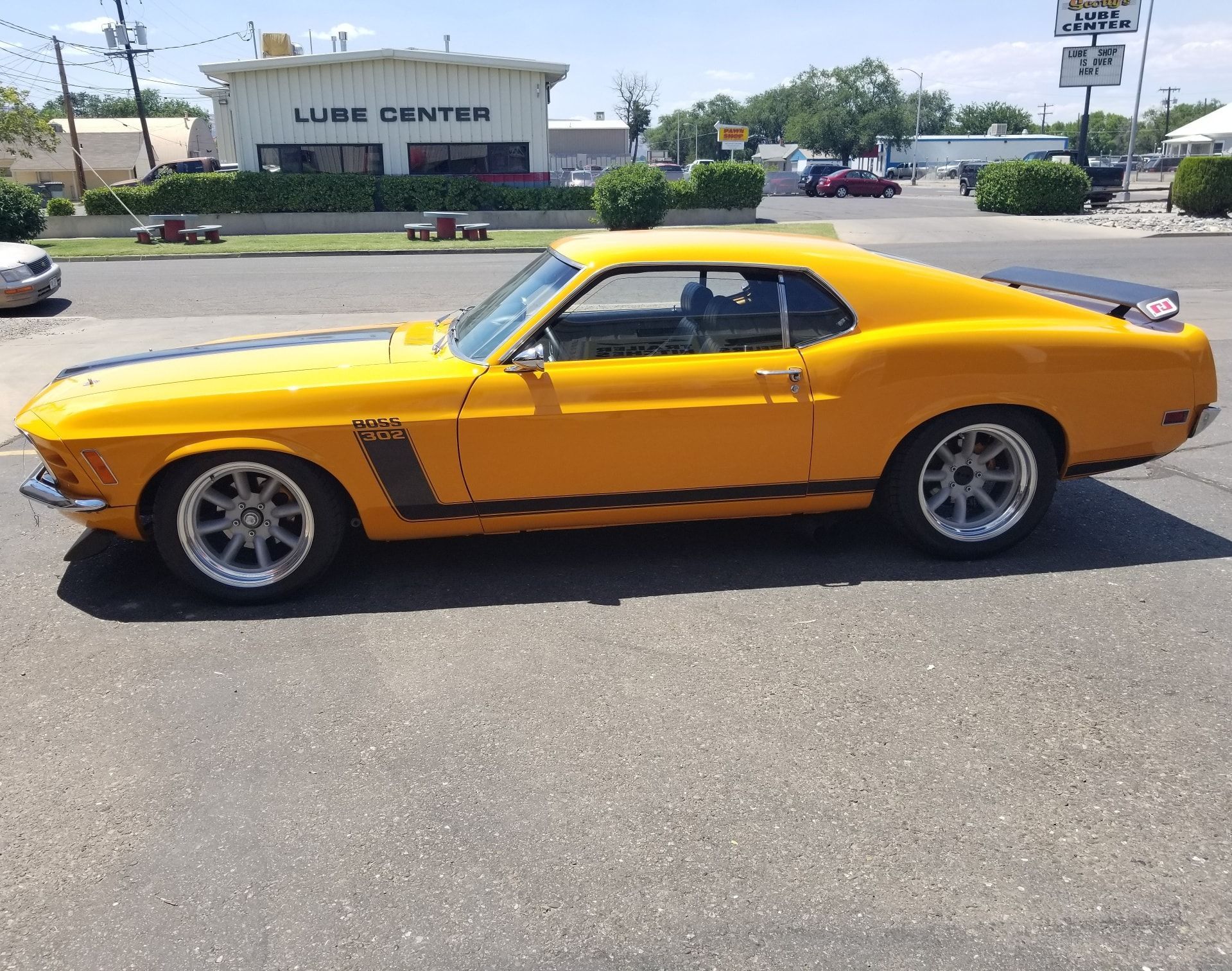 Yellow vintage Ford Mustang Boss 302 parked on a sunny street, black stripes, gray wheels, and a Lube Center in background.