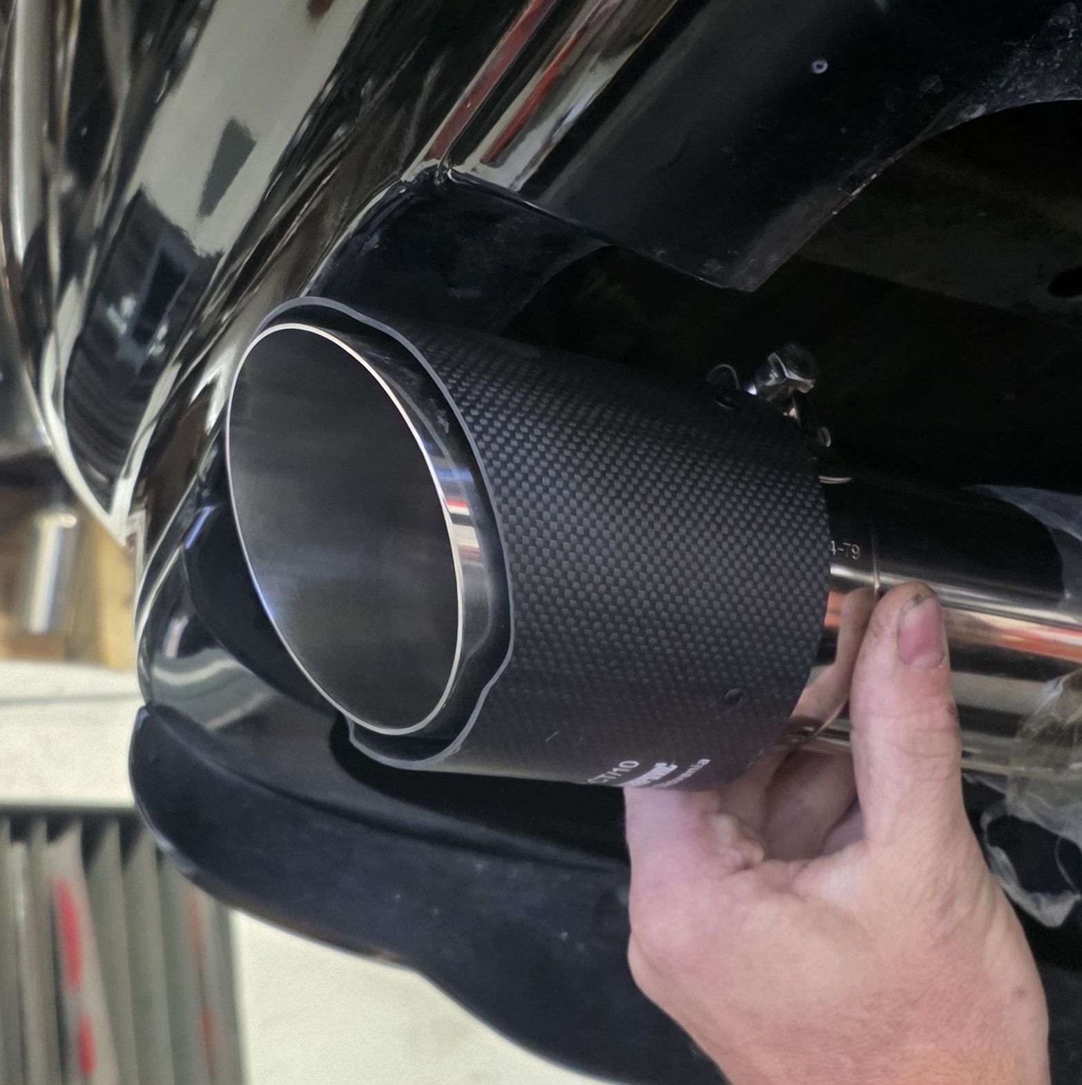 A hand attaching a carbon fiber exhaust tip to a chrome exhaust pipe on a black vehicle.