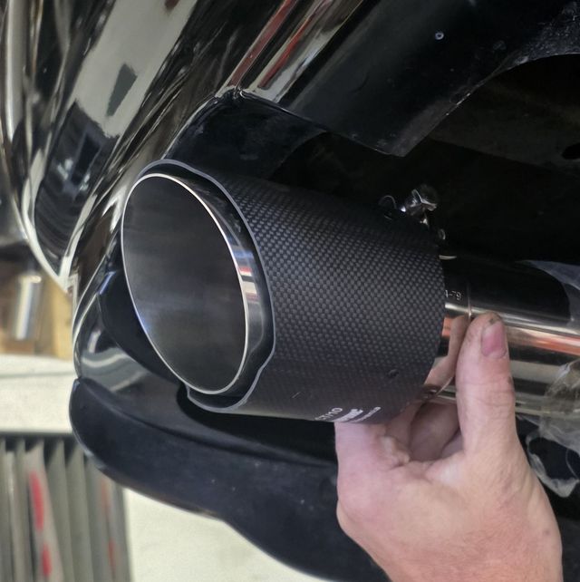 A hand attaching a carbon fiber exhaust tip to a chrome exhaust pipe on a black vehicle.