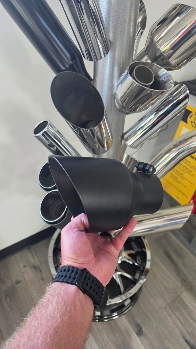 Hand holding a black exhaust tip, with various chrome exhaust tips in the background.