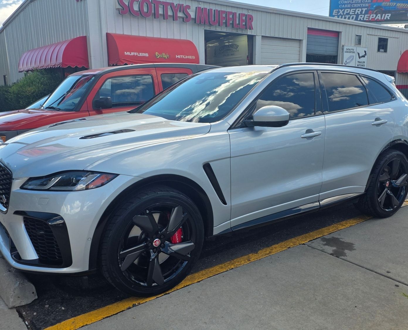 Silver Jaguar F-Pace SUV parked outside a muffler shop, with black wheels, tinted windows, and red brake calipers.