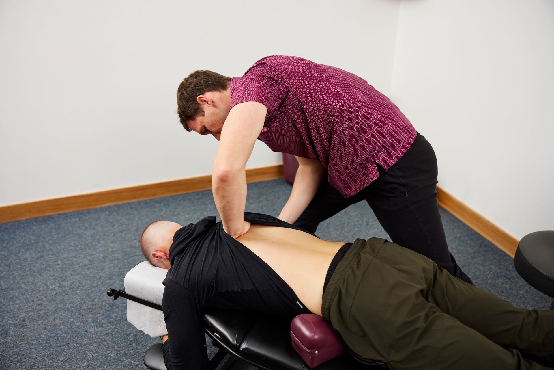 Person adjusting a reclining client on a massage table in a room with blue carpet and white walls.
