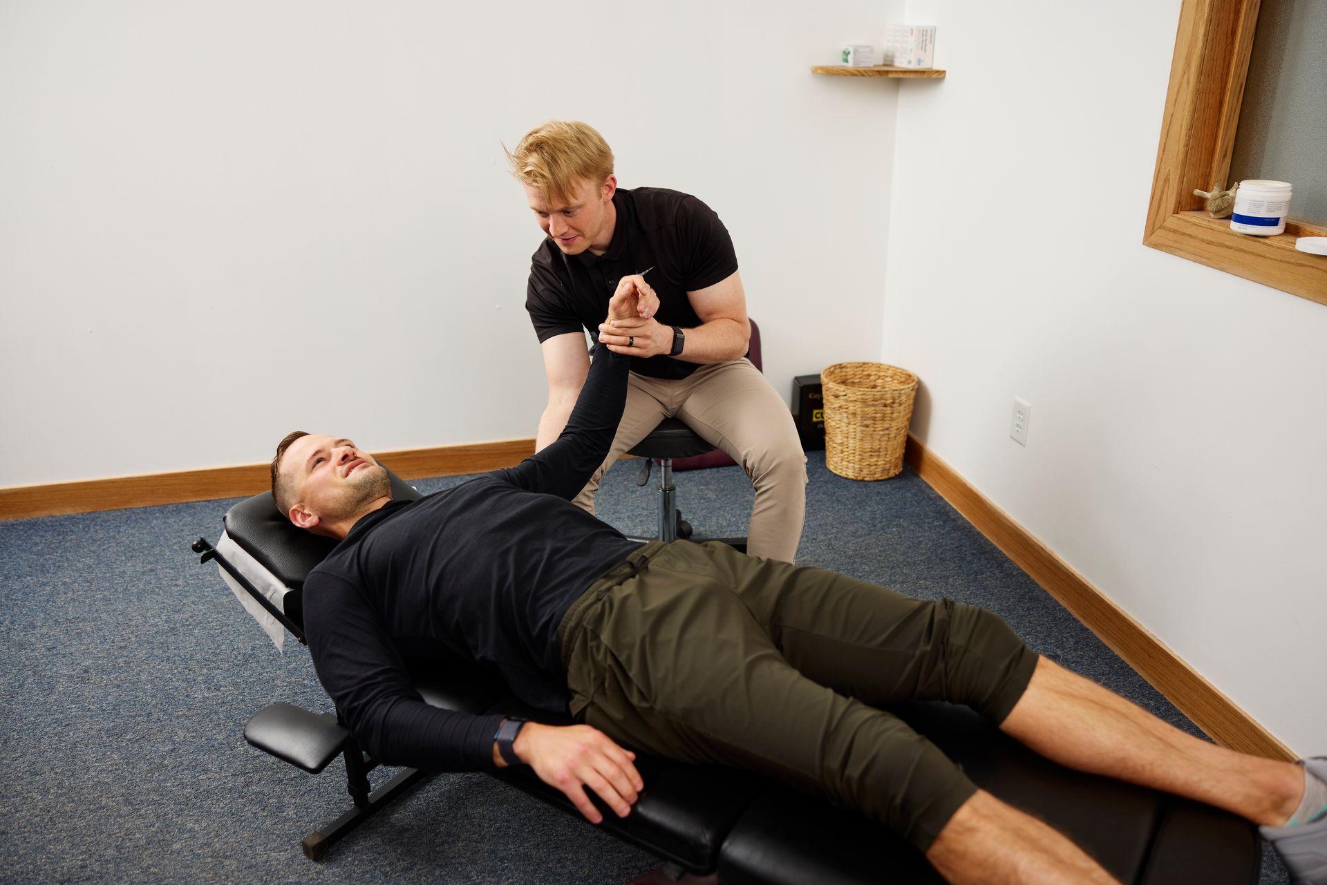 Two people in a therapy room, one assisting the other on a black treatment table.
