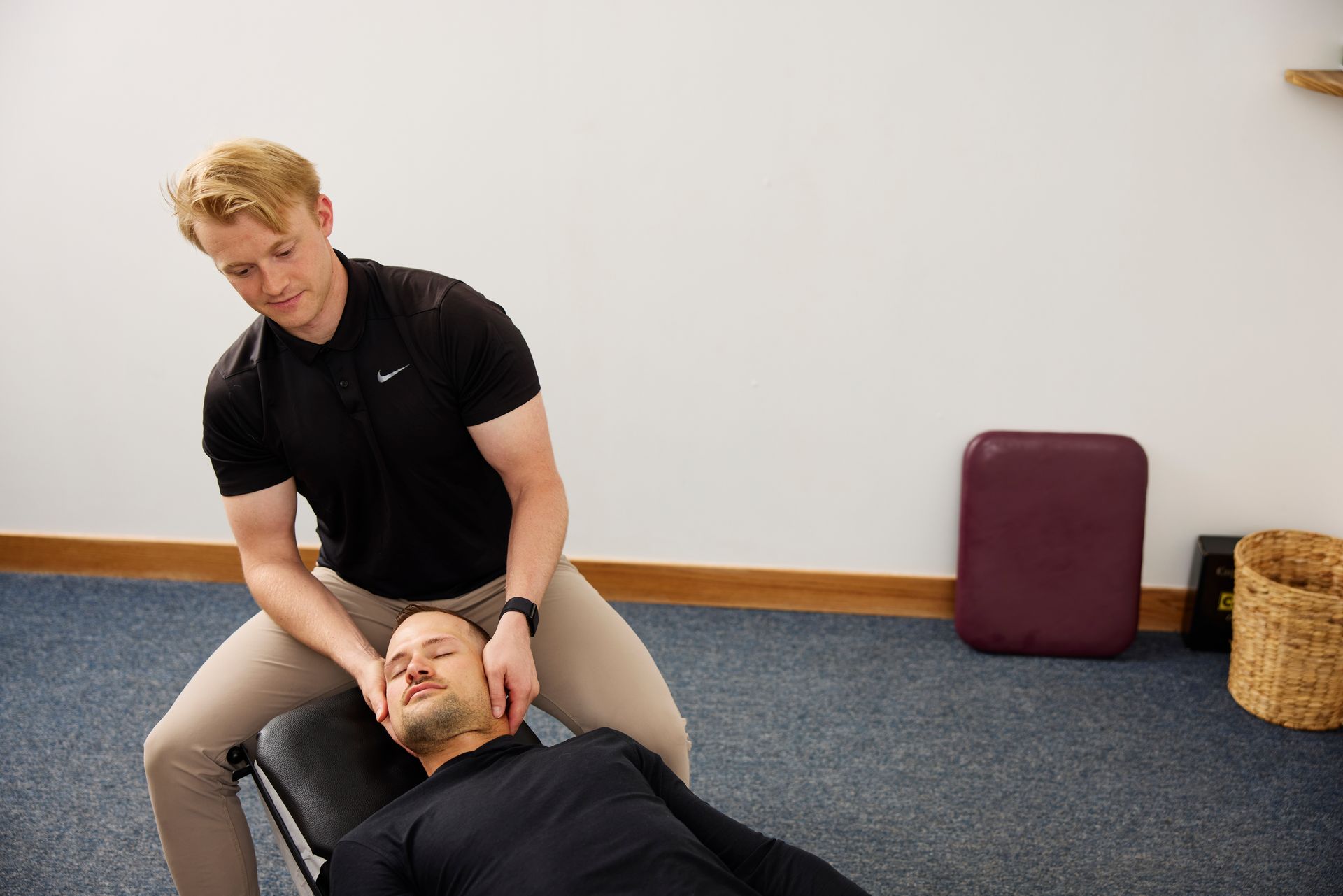 Person assisting another lying on the floor in a room with blue carpet and a plain wall.