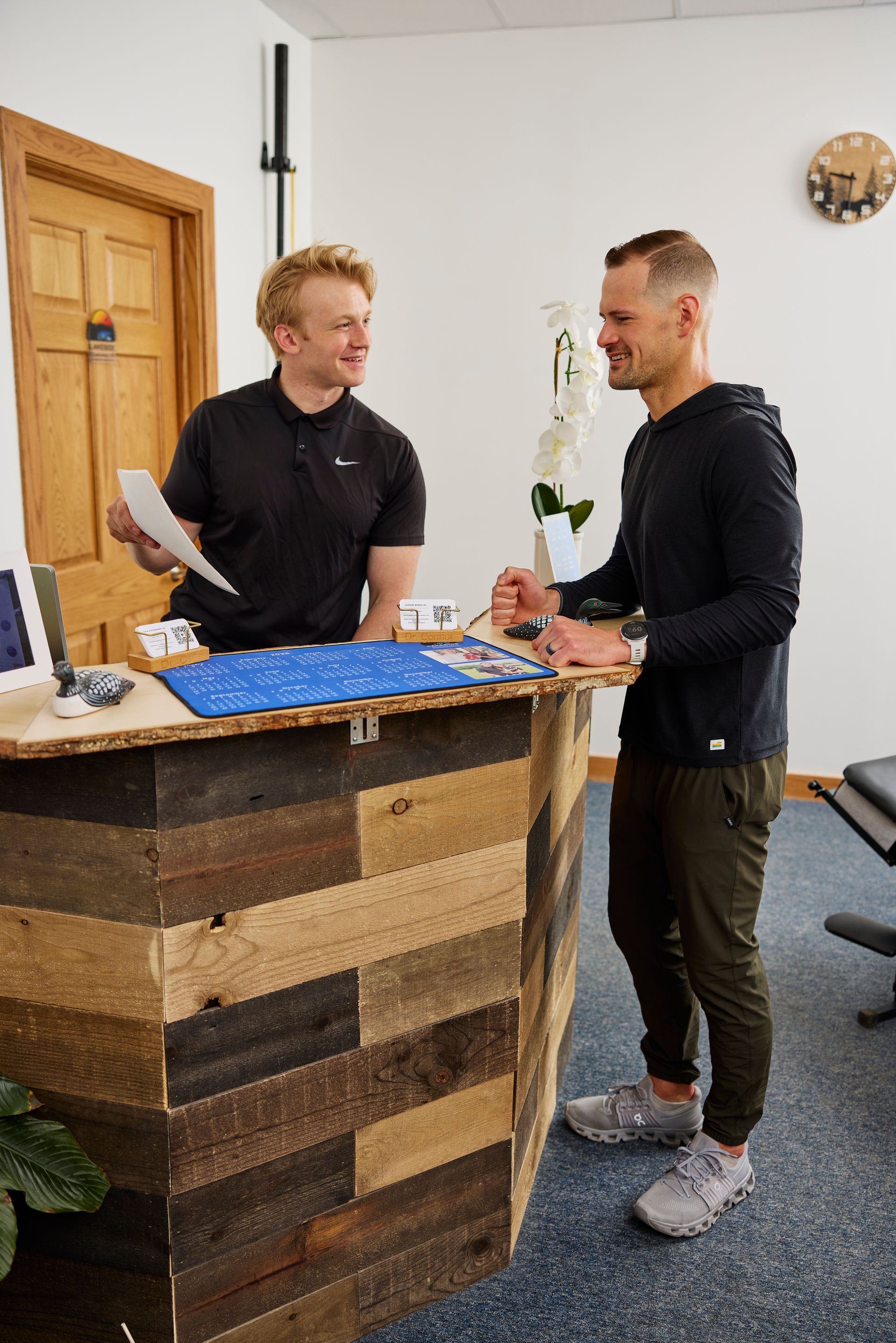 Two people smiling at a rustic reception desk, talking beside a blue tray and flowers.