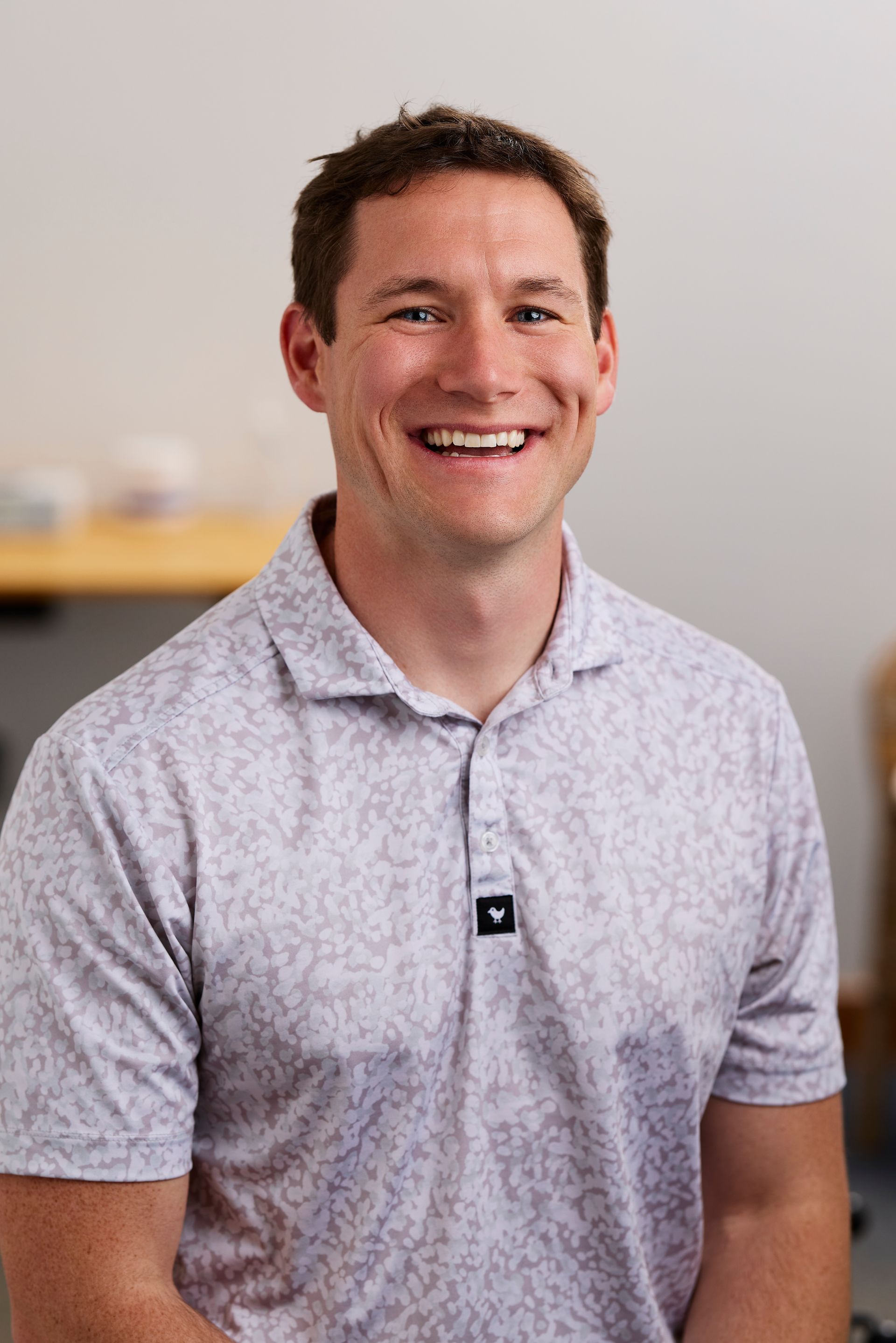 Smiling man in a light patterned polo shirt standing indoors with a blurred office background.