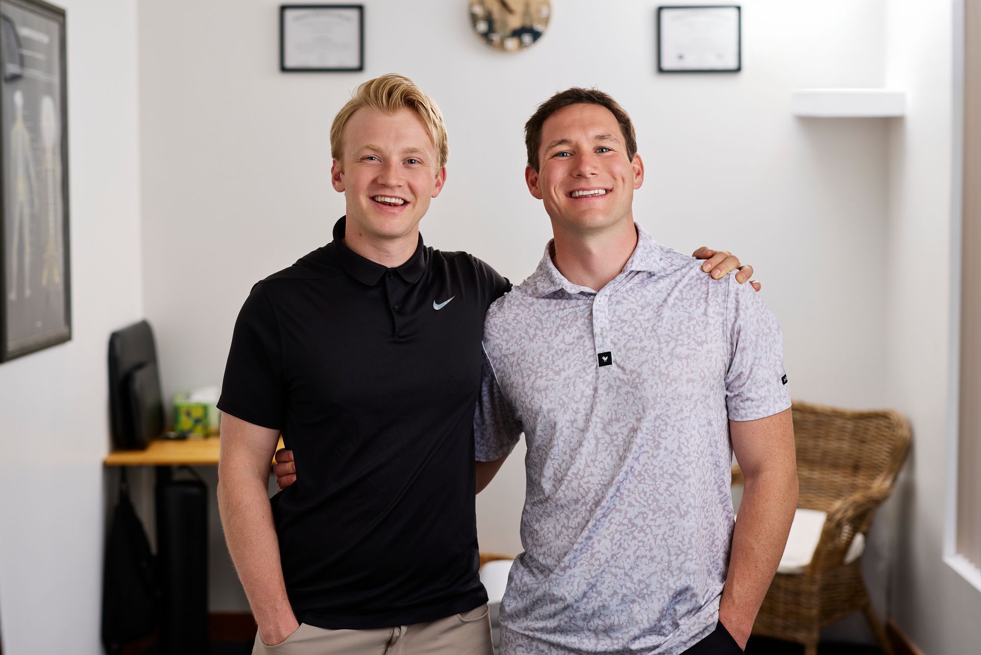 Two smiling people standing together in a bright room with framed wall art and a shelf in the background.