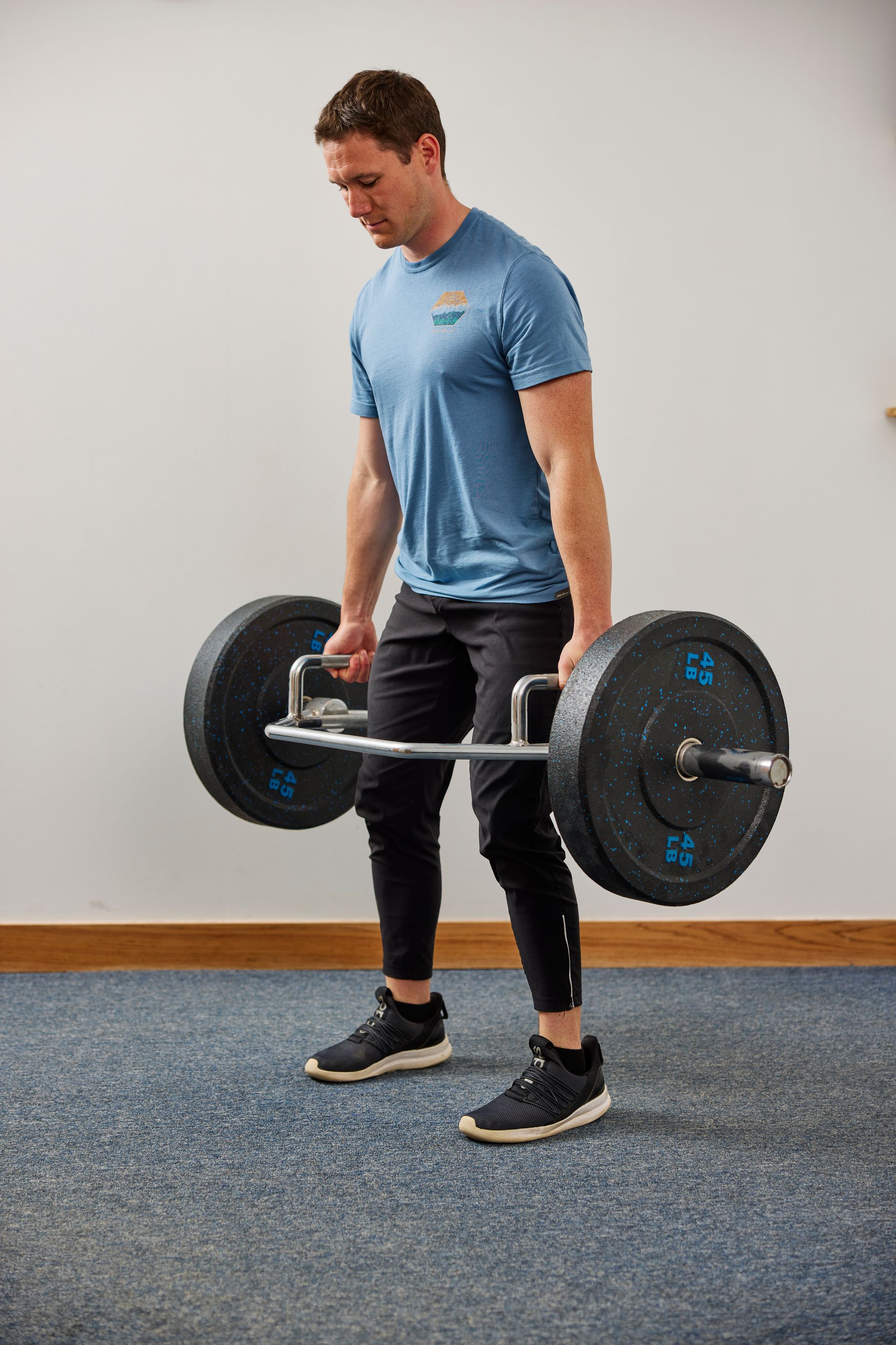 Man lifting a barbell in a gym, wearing a blue shirt and black leggings.