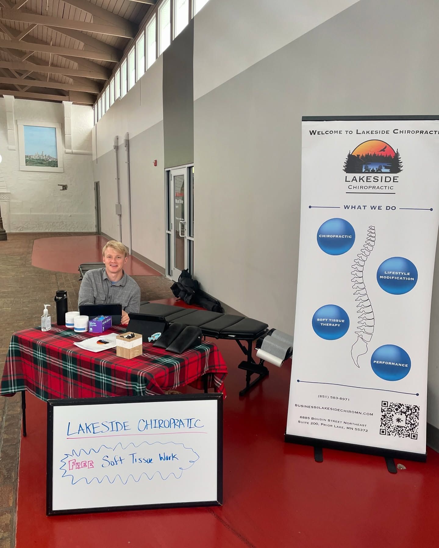 A person sits at a Lakeside Chiropractic table in a hallway next to a vertical banner listing services and a sign.