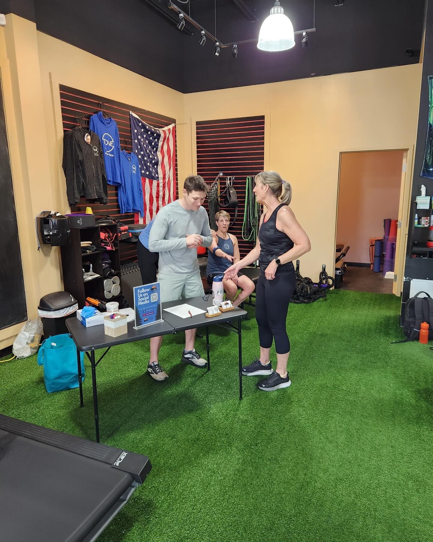 Two people stand at a table in a gym with green turf flooring, while another person sits behind them in the background.