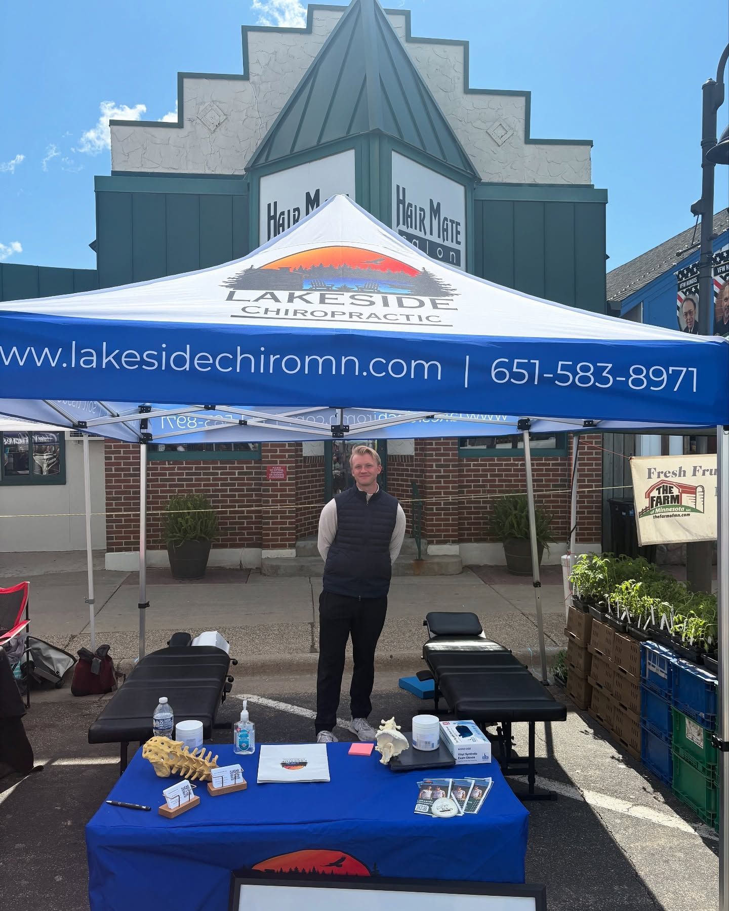 A person stands under a Lakeside Chiropractic tent at an outdoor market with a table of medical models and supplies.