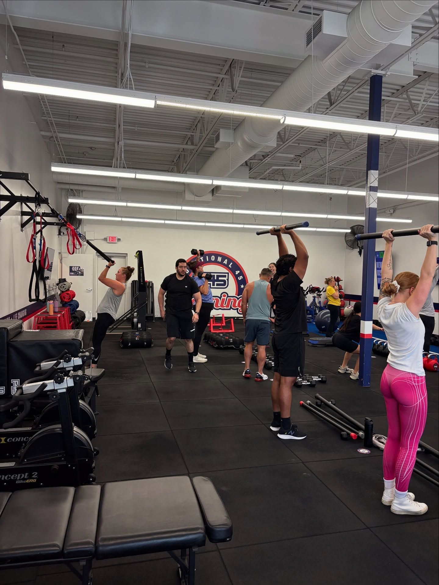 People in a gym performing overhead barbell lifts and other exercises in a well-lit fitness studio.