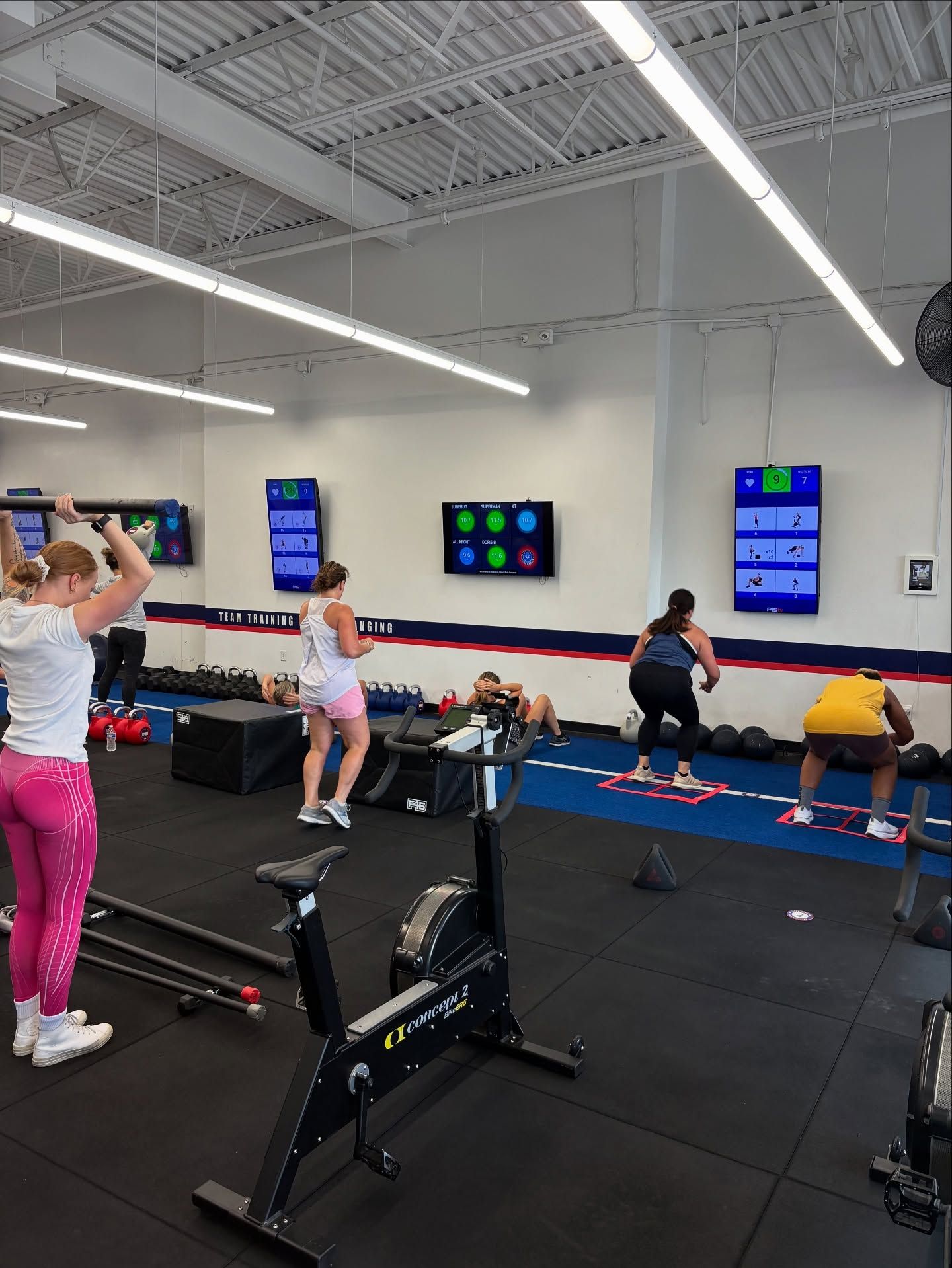 Four people perform various exercises in a brightly lit fitness studio equipped with monitors and workout equipment.