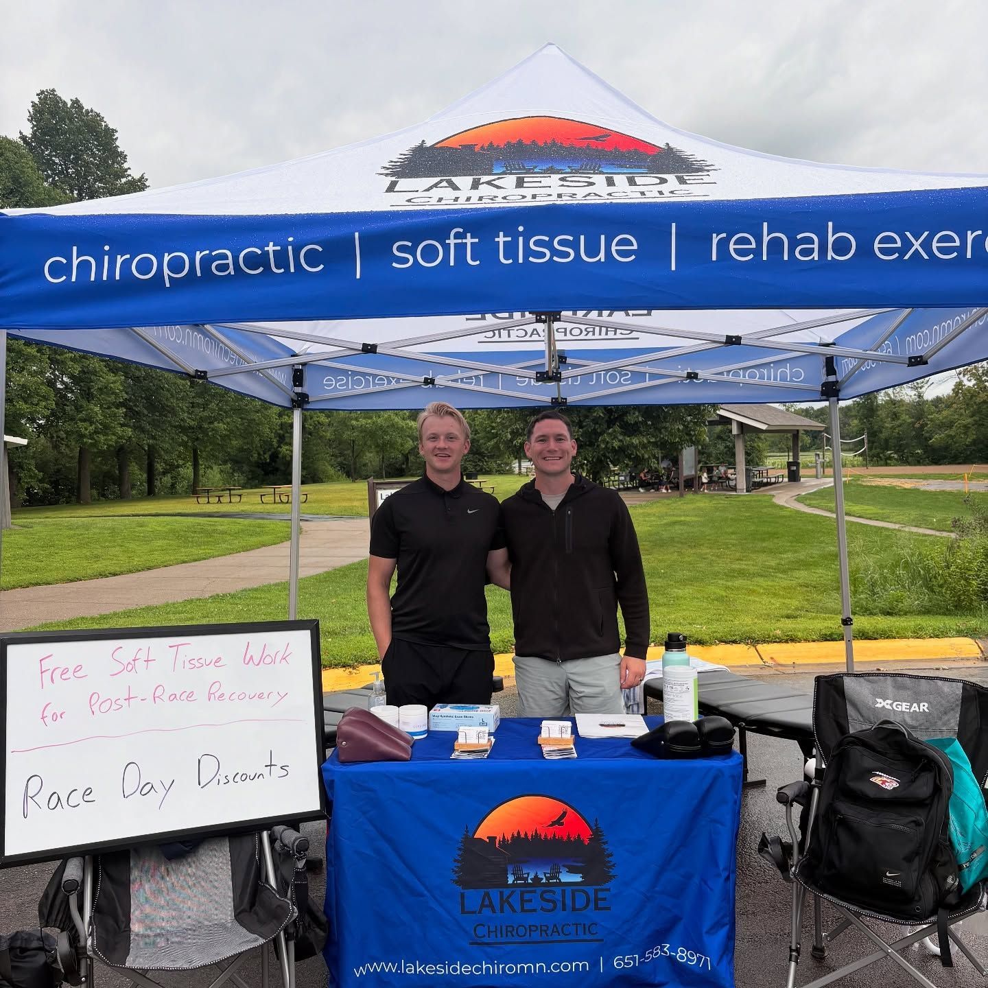 Two people stand at a Lakeside Chiropractic event booth outdoors with a sign offering soft tissue work for race recovery.