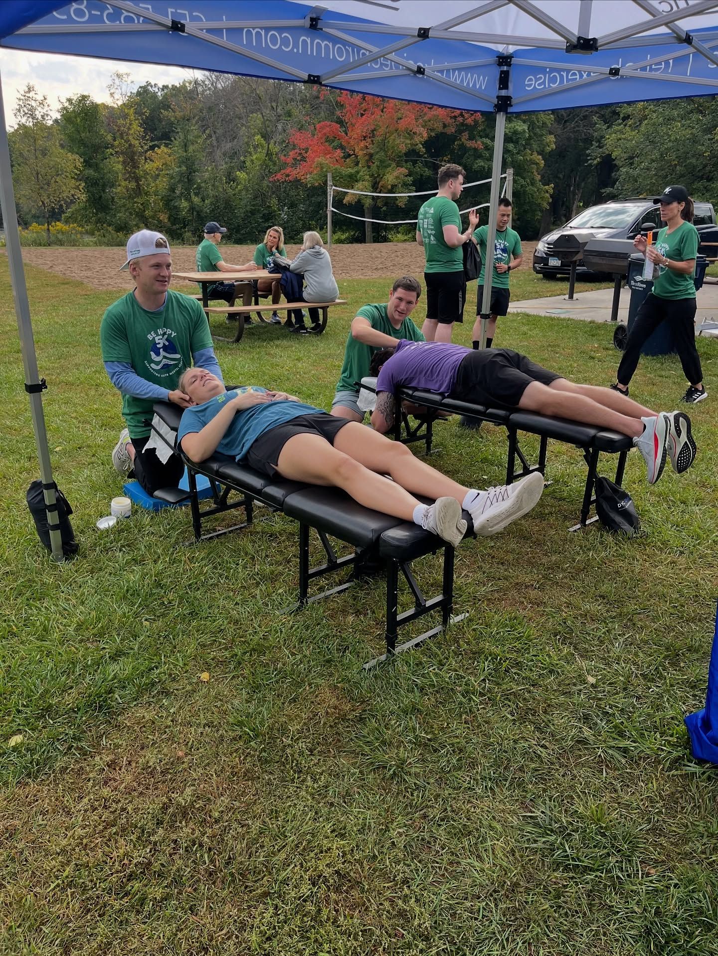 People receive sports massage treatments on portable tables under a blue tent at an outdoor athletic event.