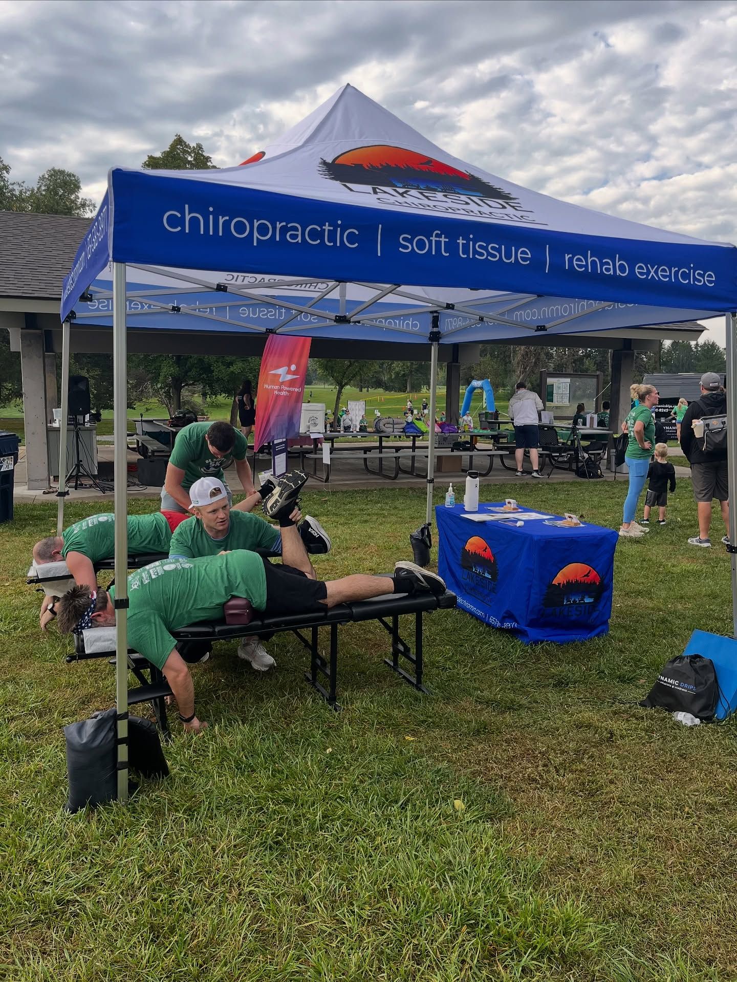 People in green shirts receive chiropractic treatment at a pop-up tent in a park.