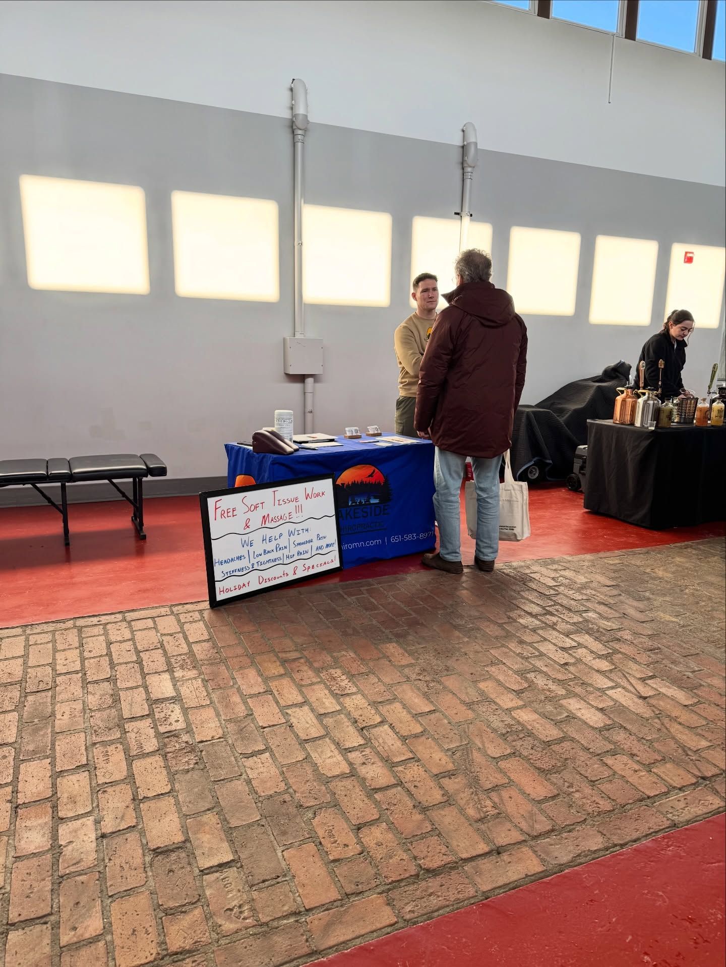 People stand at a trade show table with a sign in a large hall with brick flooring and bright rectangular window panels.