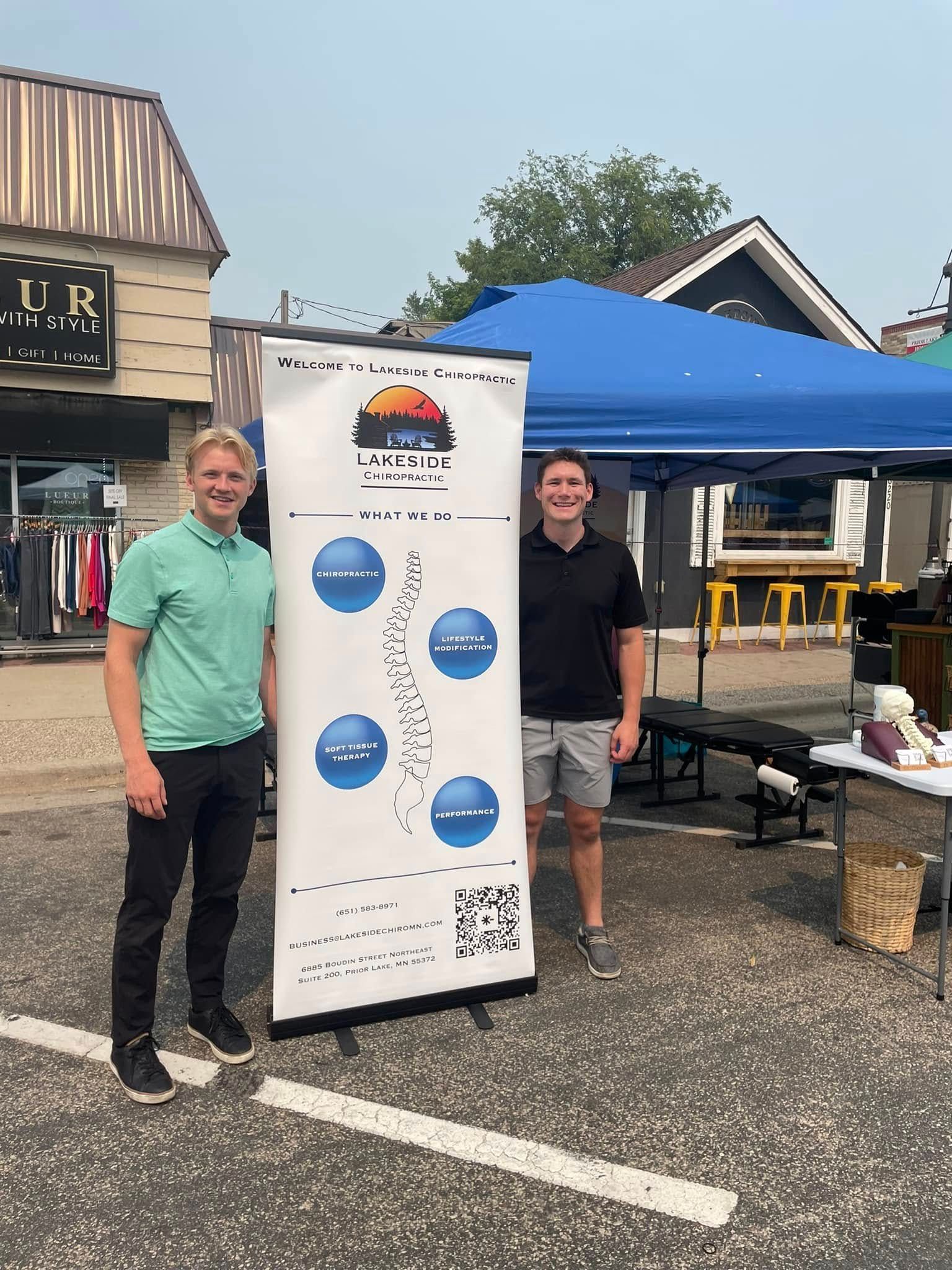 Two people stand outdoors next to a tall banner for a chiropractic business, with a blue tent in the background.