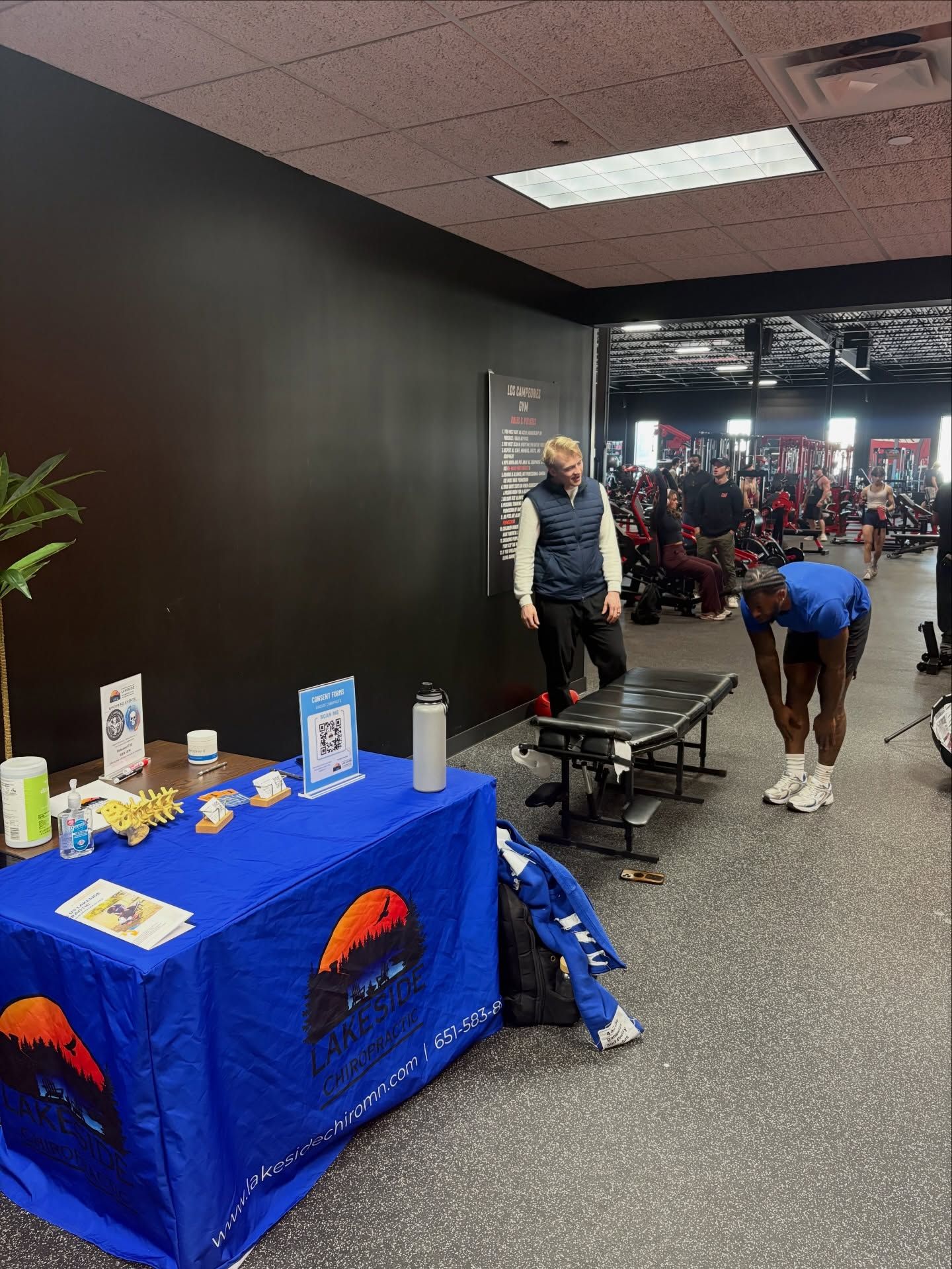 A blue promotional table sits in a gym hallway, while two people stand nearby, one stretching and one standing by a bench.