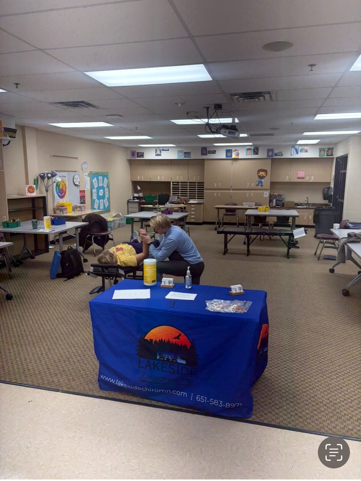 A person in a blue shirt sits at a table in a carpeted room, assisting someone lying on a massage table.