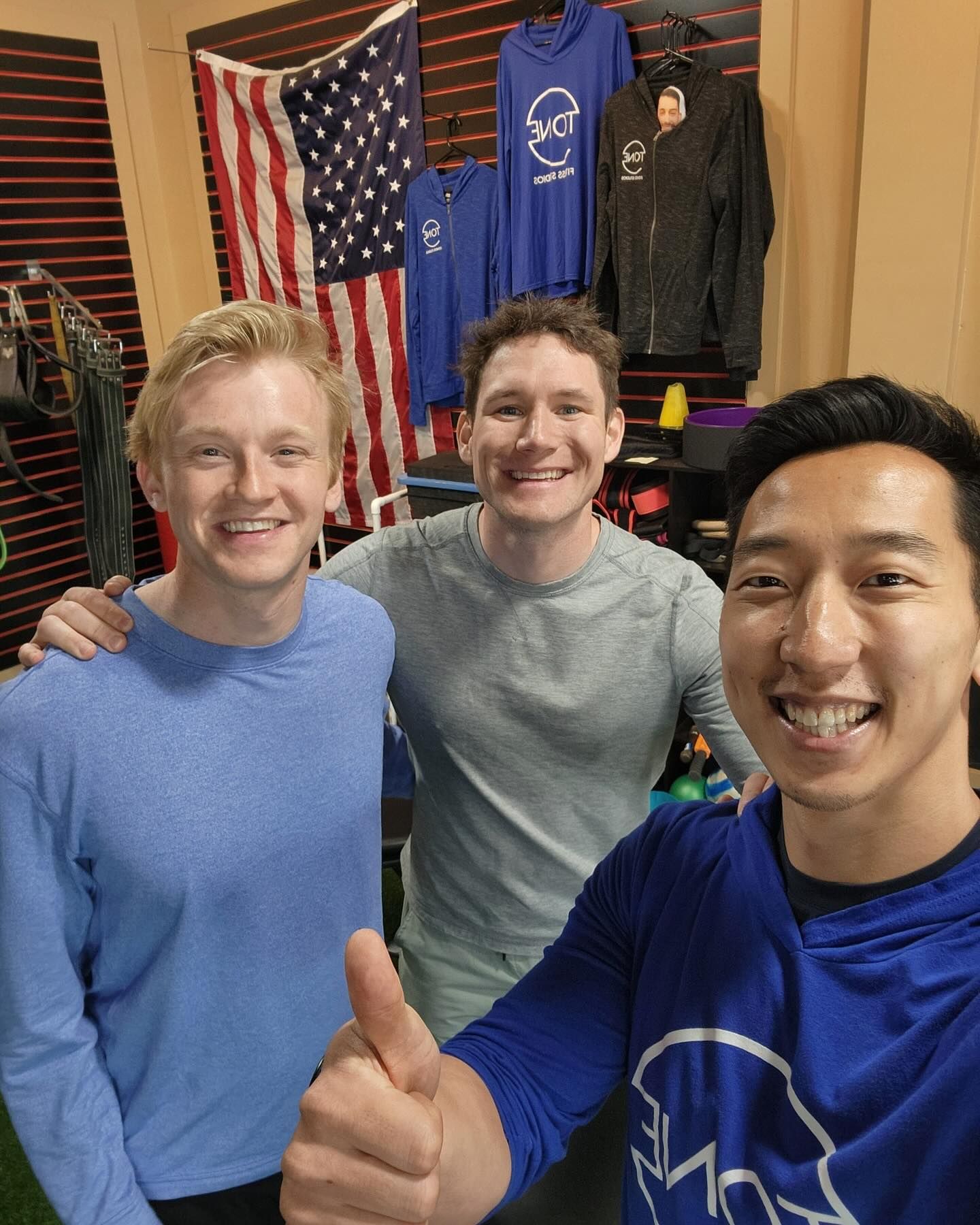 Three smiling people pose for a selfie in a gym with an American flag and hanging workout gear in the background.