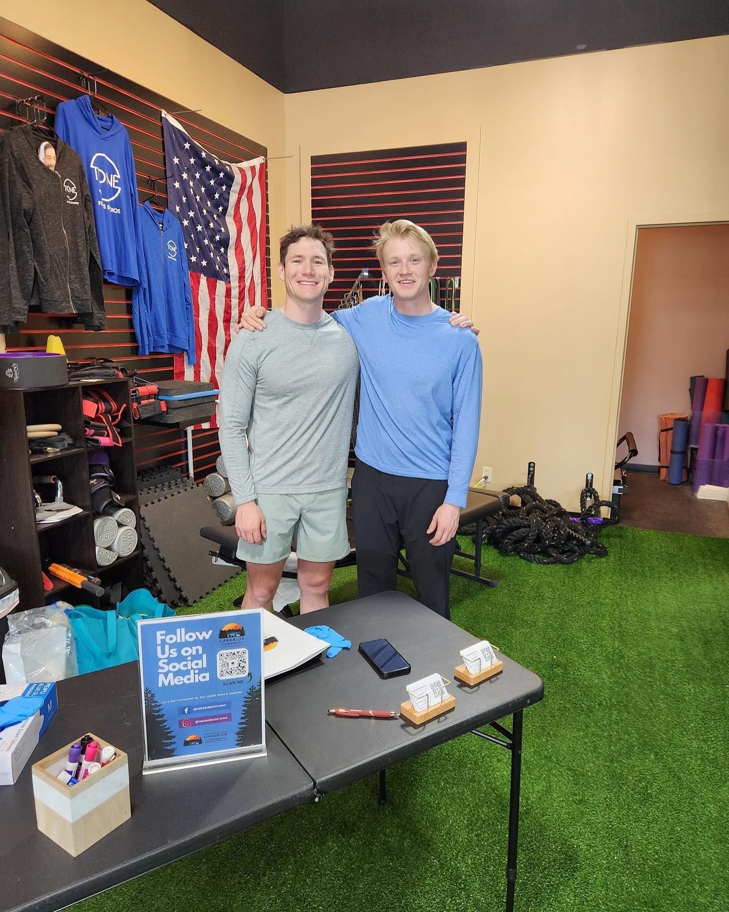 Two smiling people stand behind a table on green turf in a shop with hanging athletic gear and a US flag in the background.