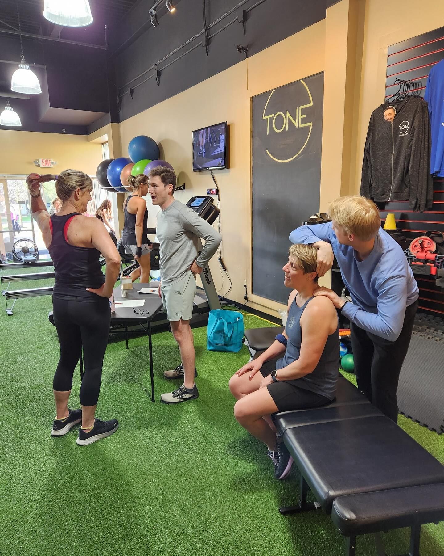 A fitness professional assists a person sitting on a gym bench, while others stand and chat in a bright indoor studio.