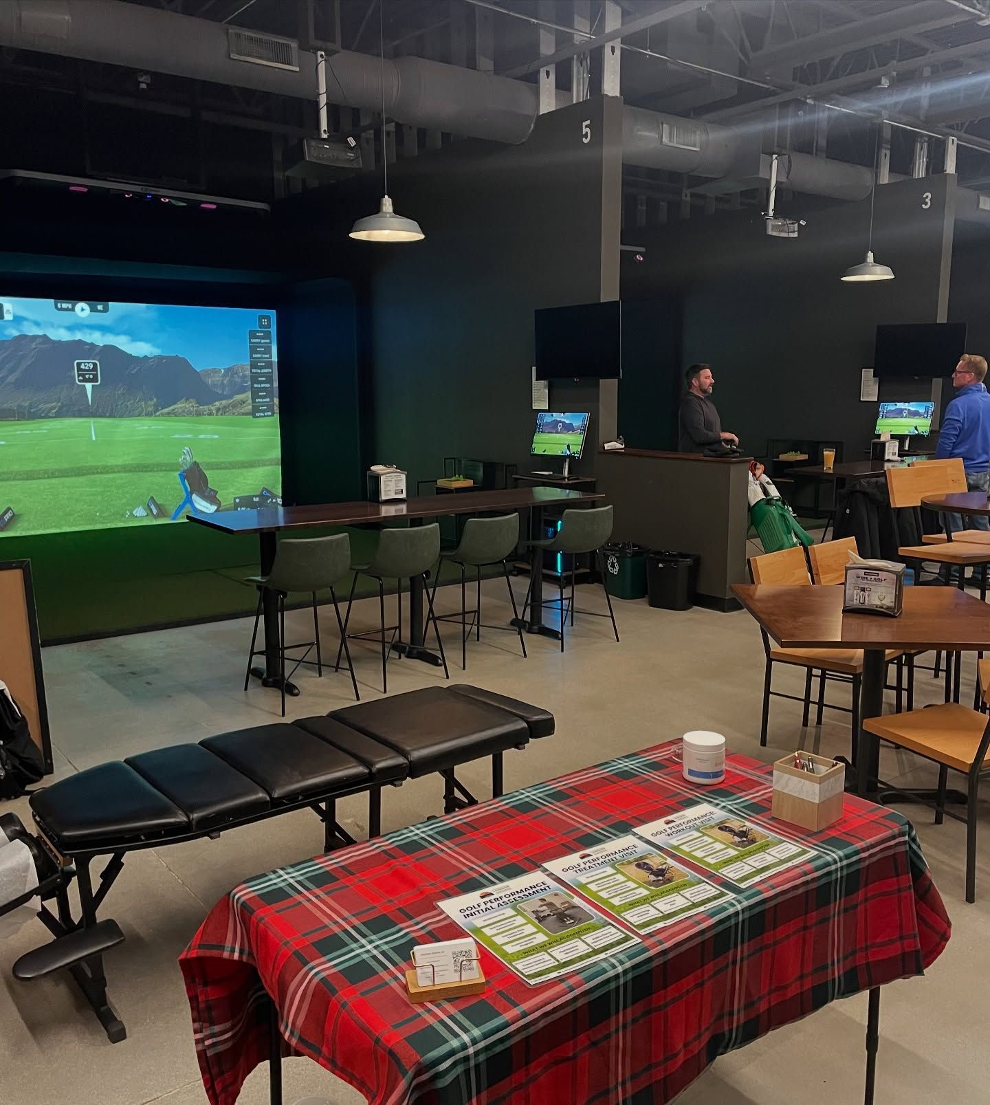 An indoor golf simulator facility with green screens, bar seating, and a table with a red plaid tablecloth in the foreground.