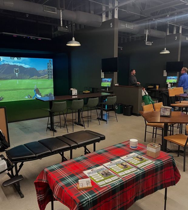 Indoor golf simulator center with a red plaid table in the foreground, dining tables, and a large projection screen.