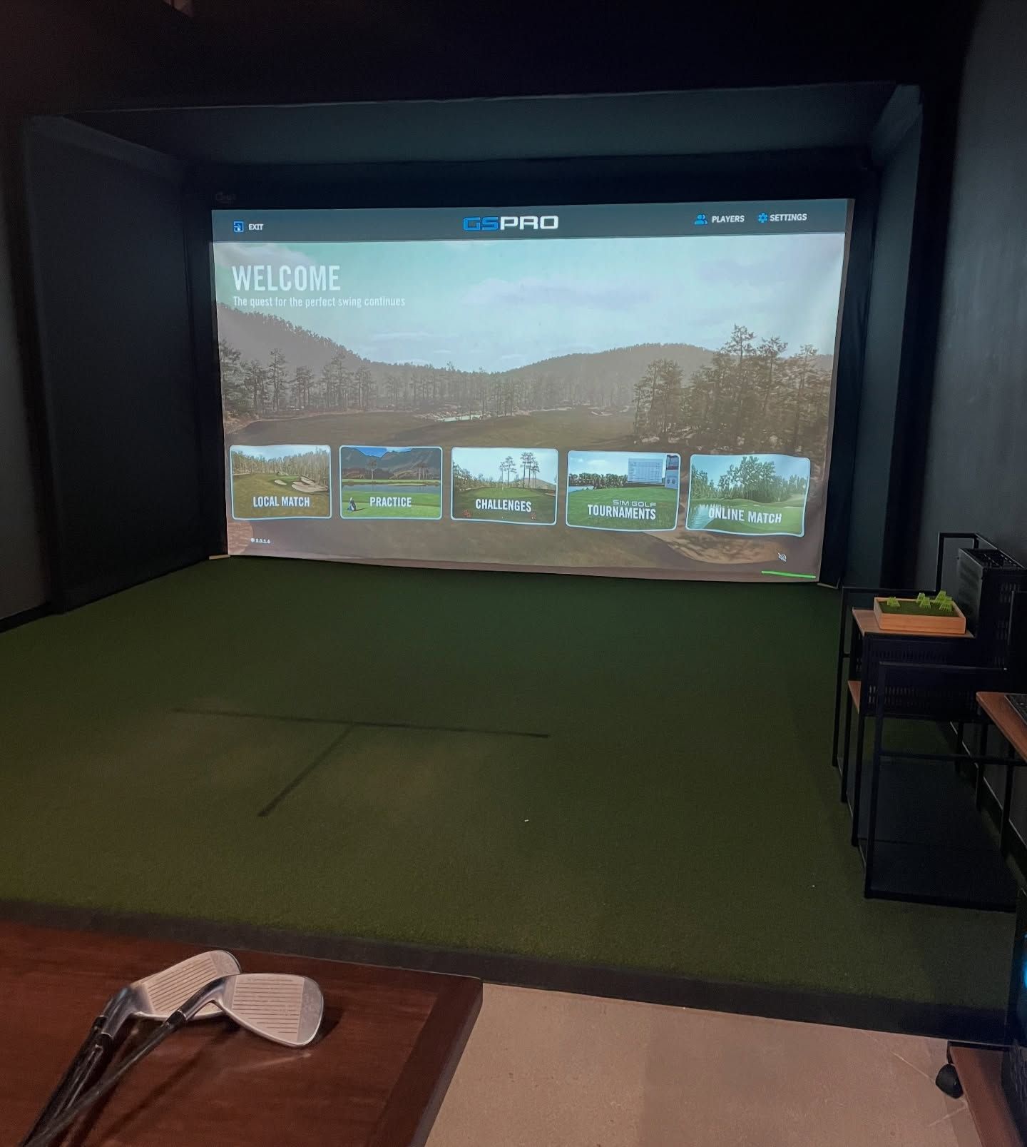 An indoor golf simulator room with a large screen displaying course options, a green turf floor, and two golf clubs.