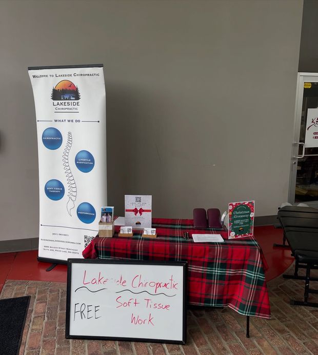 A Lake de Chiropractic booth with a branded banner, a plaid-covered table, and a sign offering free soft tissue work.