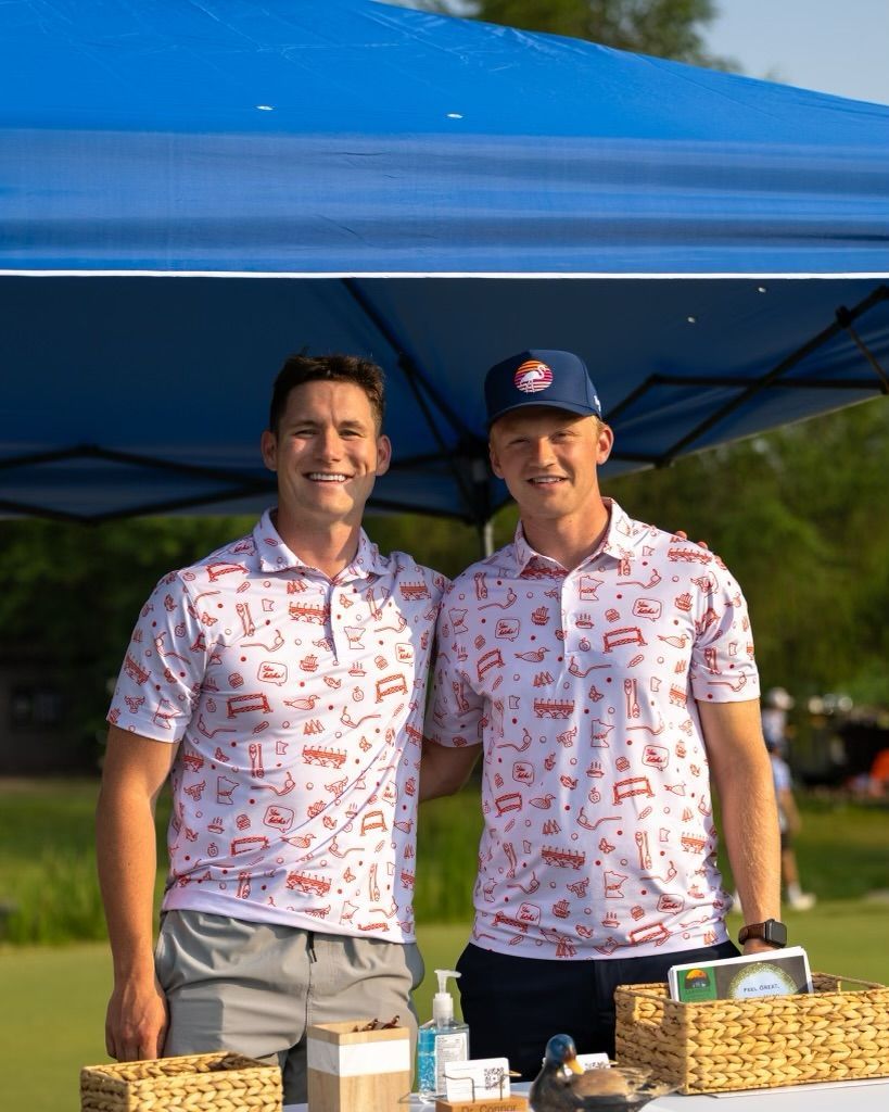 Two people stand smiling under a blue canopy, wearing matching white patterned polo shirts at an outdoor event.