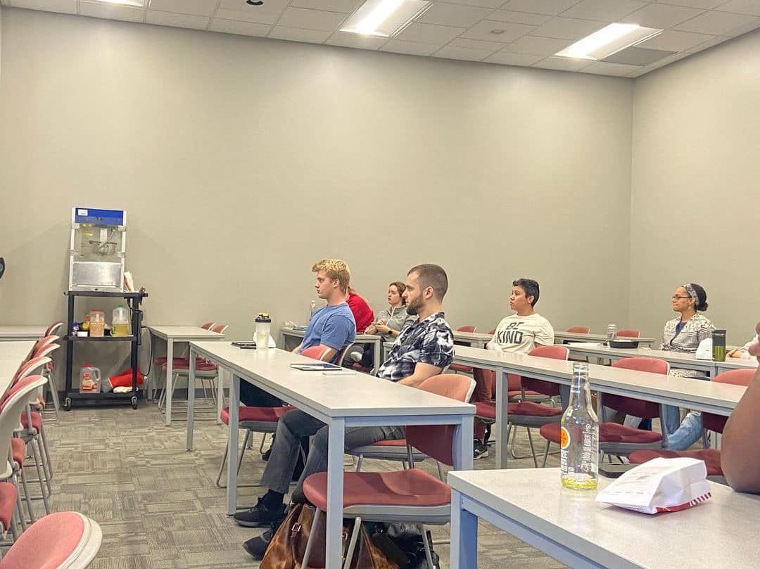 People sitting in rows at desks in a conference room, facing left toward a presenter or screen.