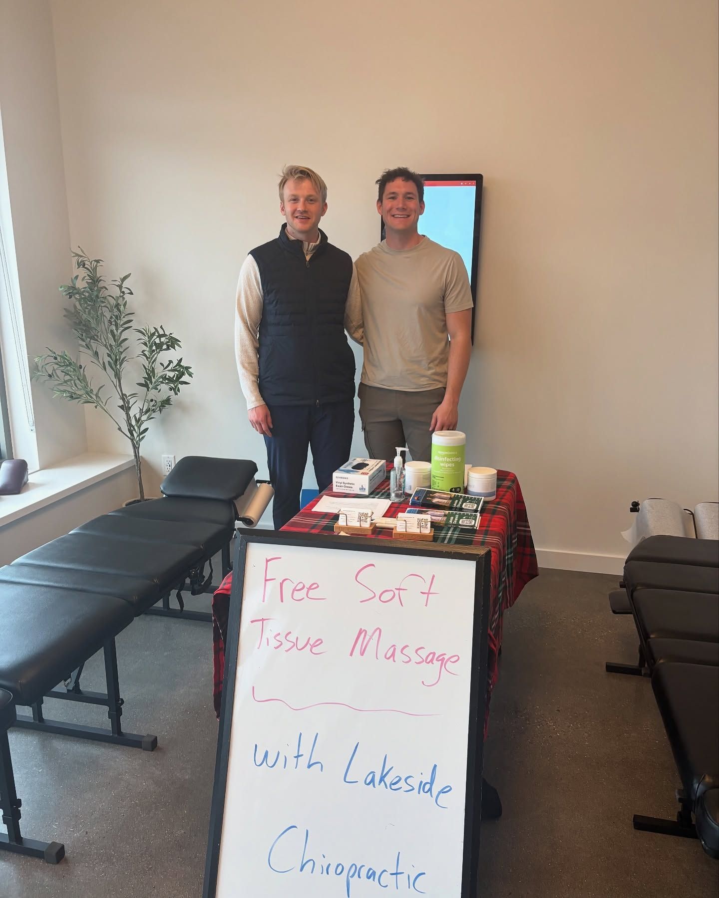 Two people stand in a clinical office behind a table with a sign advertising free soft tissue massage by Lakeside Chiropractic.