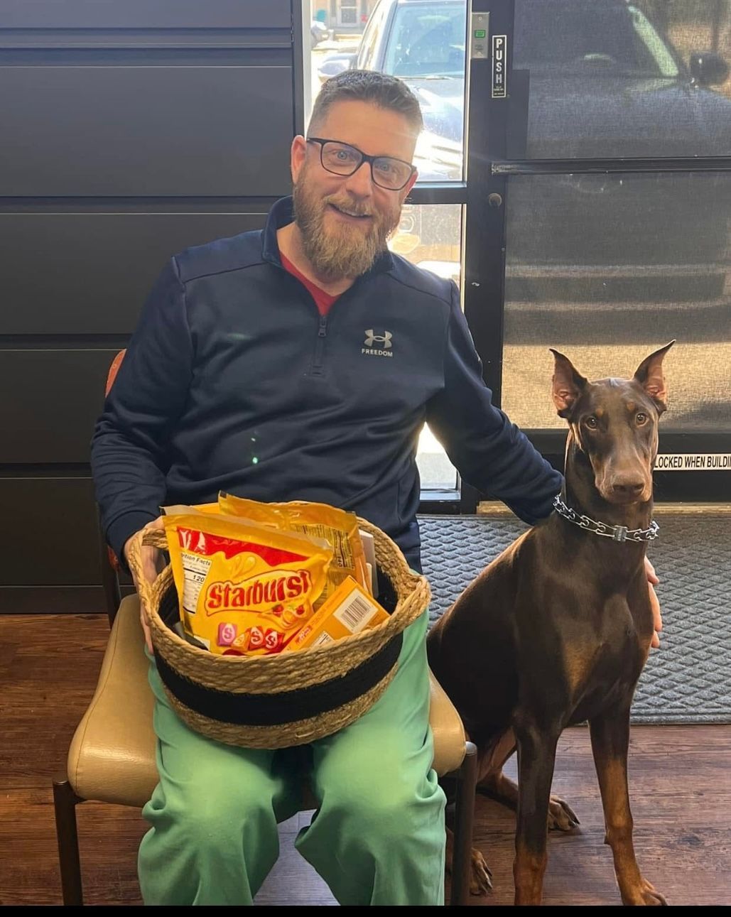 A man is sitting in a chair holding a basket of snacks next to a dog.