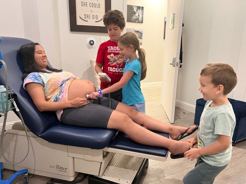 A pregnant woman is sitting in a hospital chair with two children standing around her.