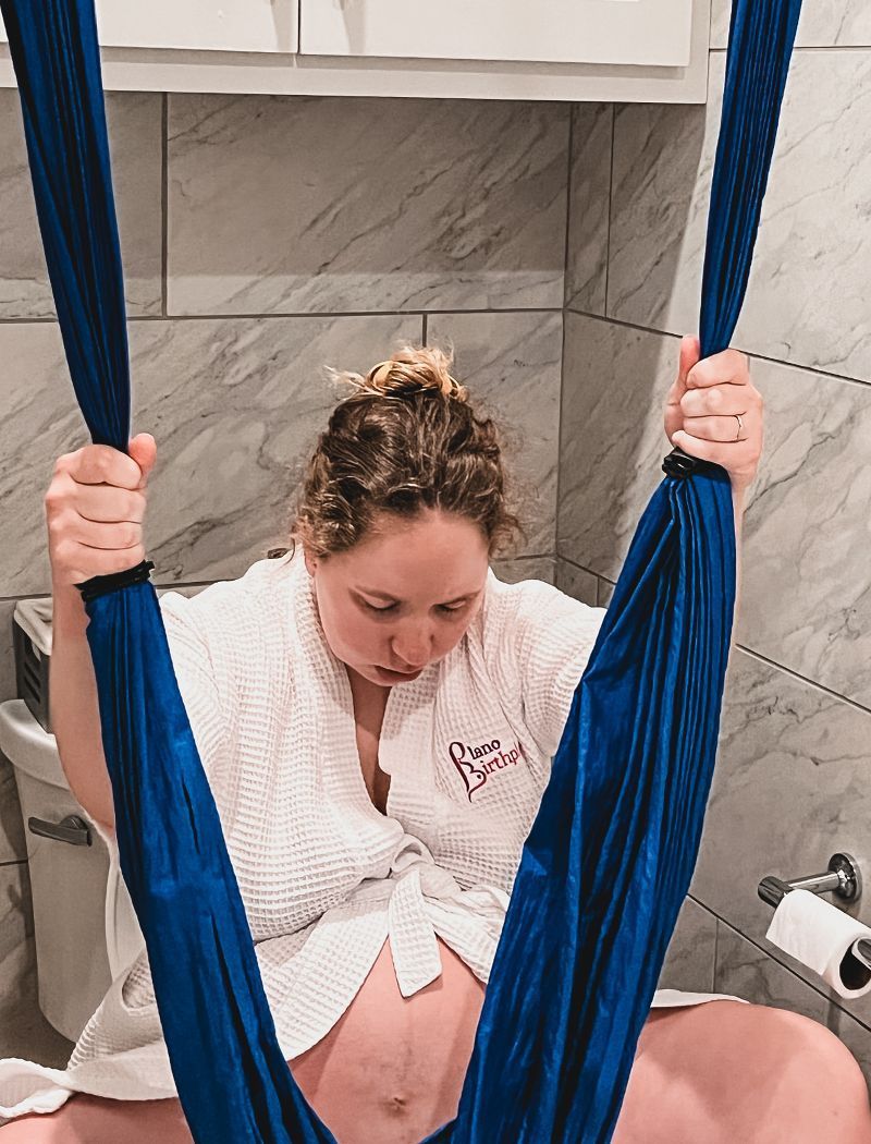 A pregnant woman is sitting in a hammock in a bathroom.