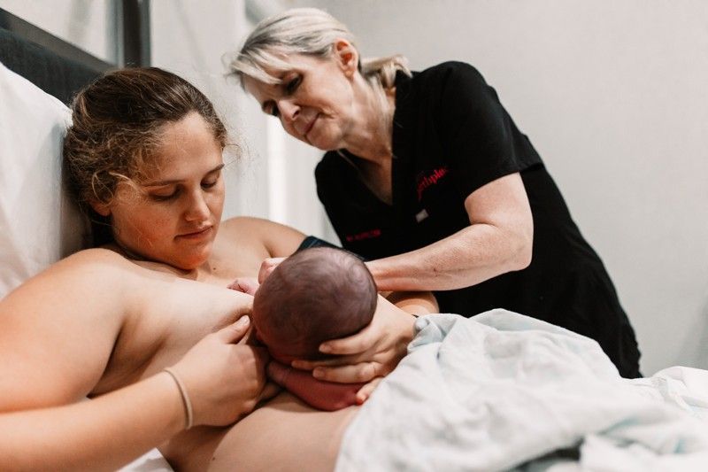 A woman is breastfeeding a baby in a hospital bed while a doctor looks on.