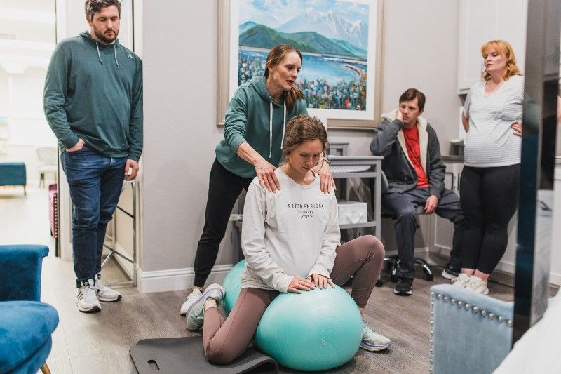 A group of people are standing around a woman sitting on a pilates ball.
