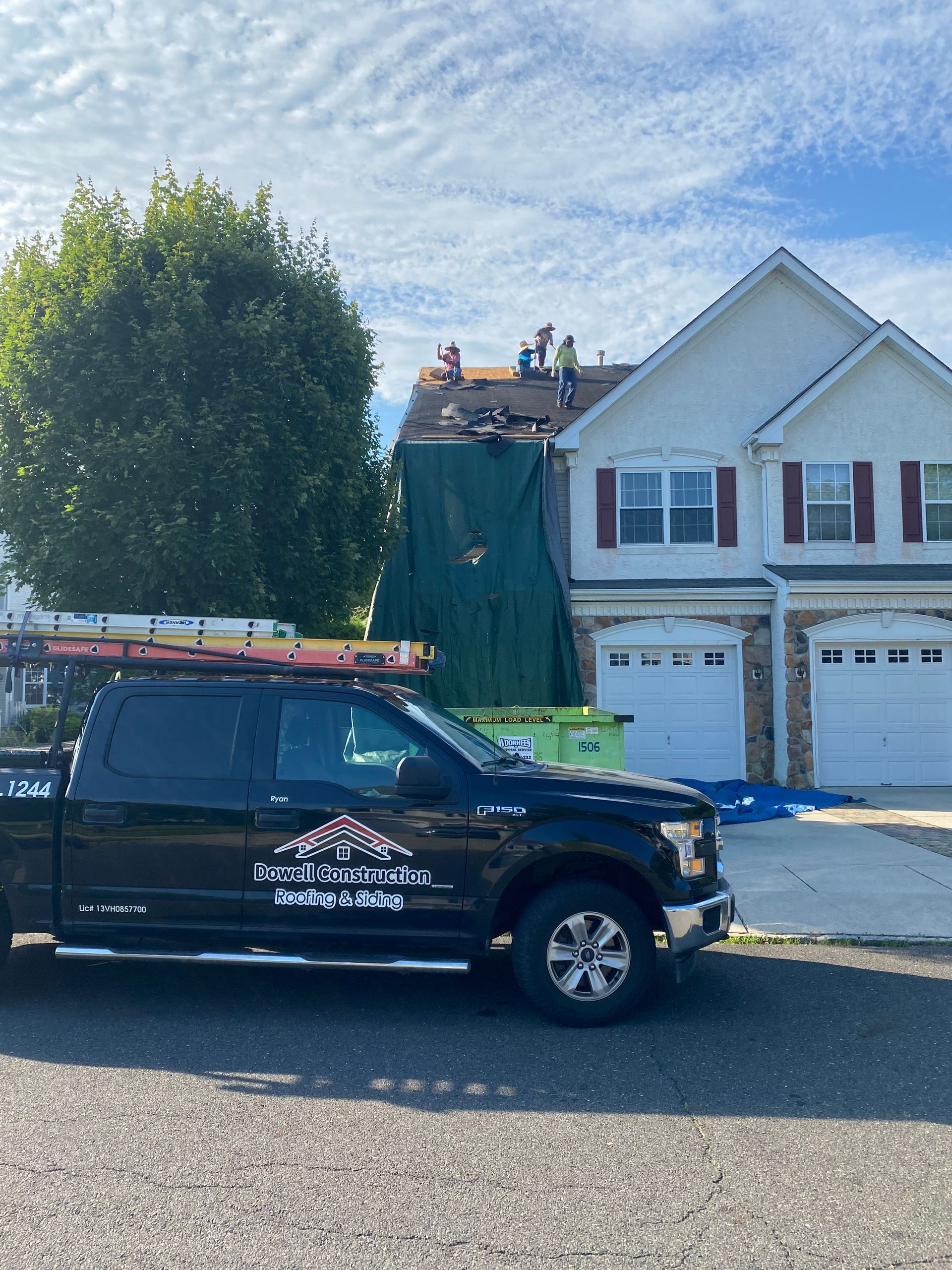 Black truck parked in front of a house with workers on the roof. Roofing supplies on the roof and a green tarp.