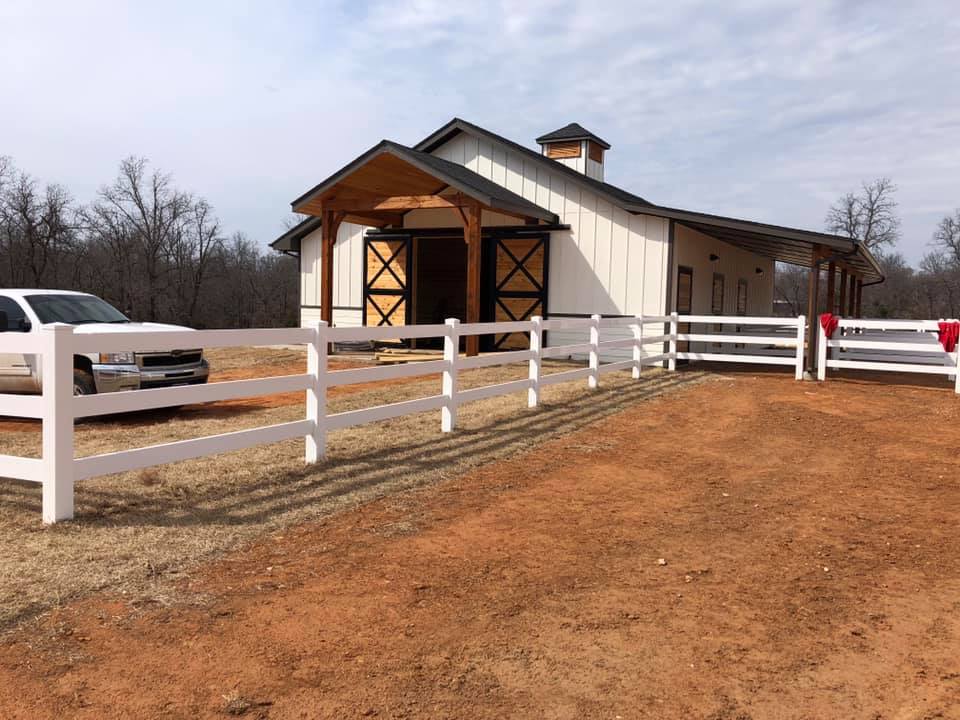 A white truck is parked in front of a barn behind a white fence.