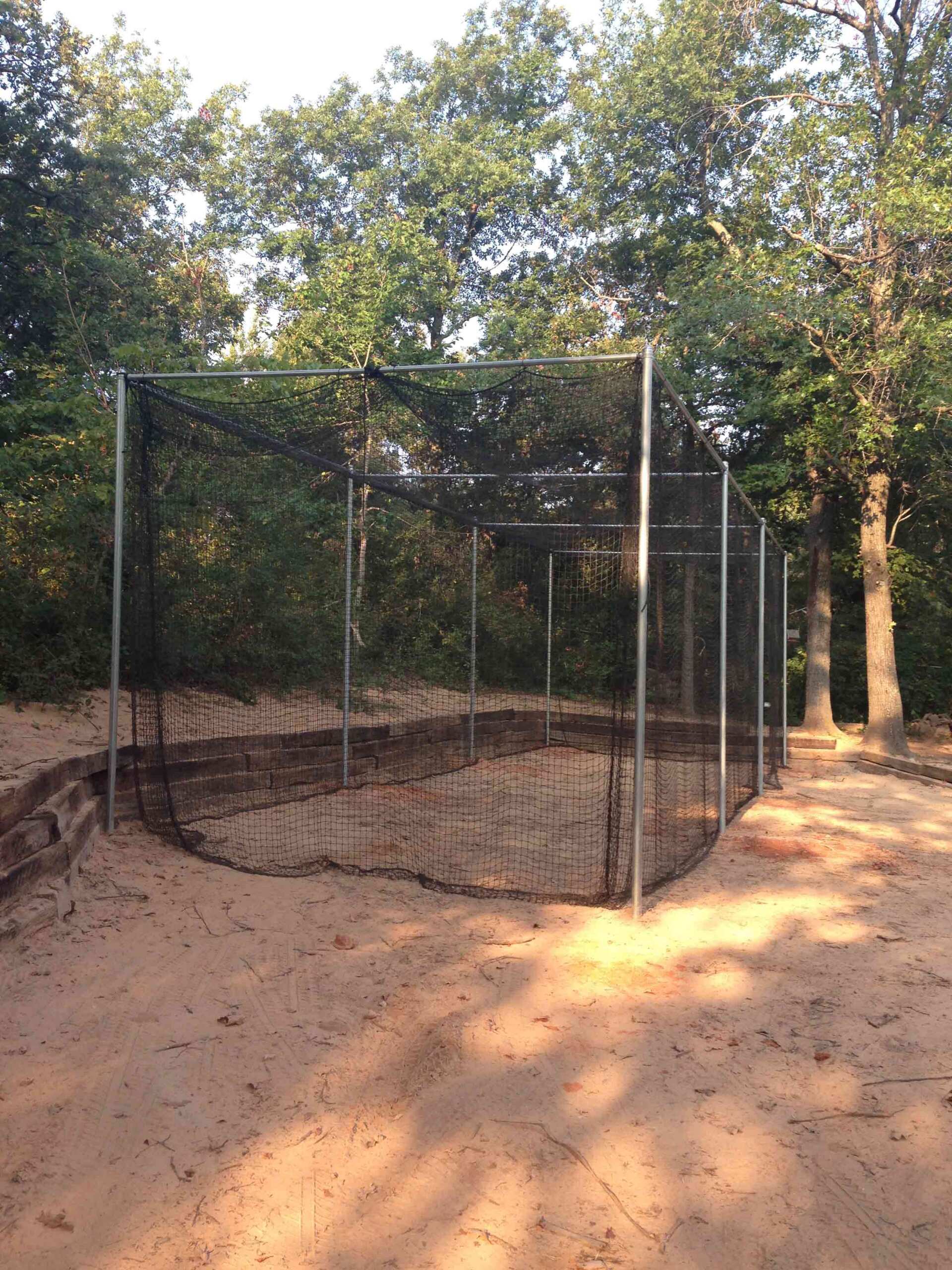 A baseball cage is sitting in the middle of a dirt field surrounded by trees.