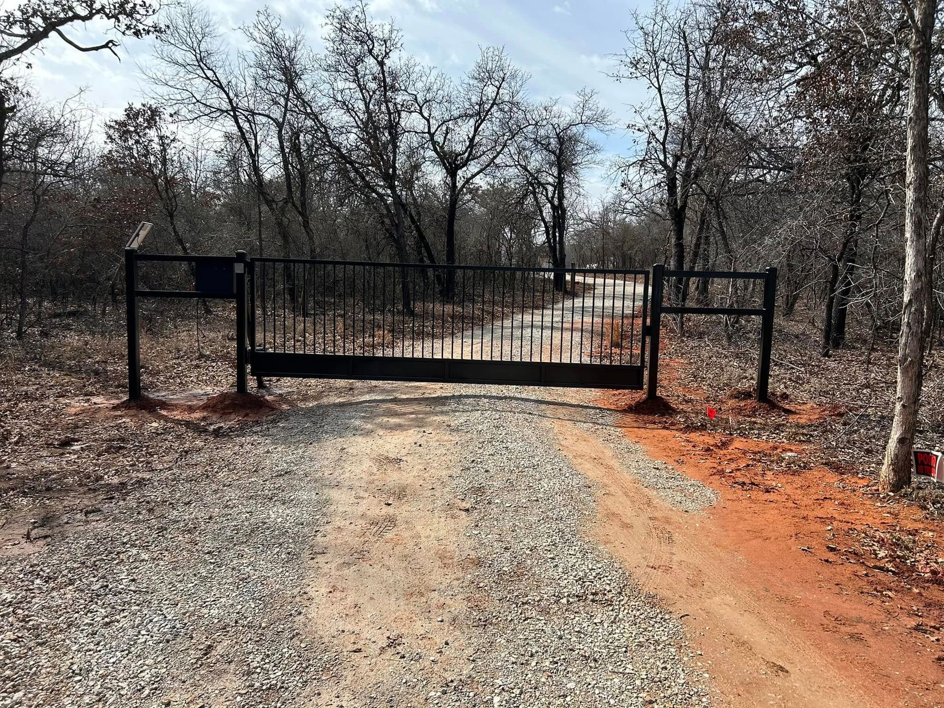 A black gate is sitting on the side of a gravel road in the middle of a forest.