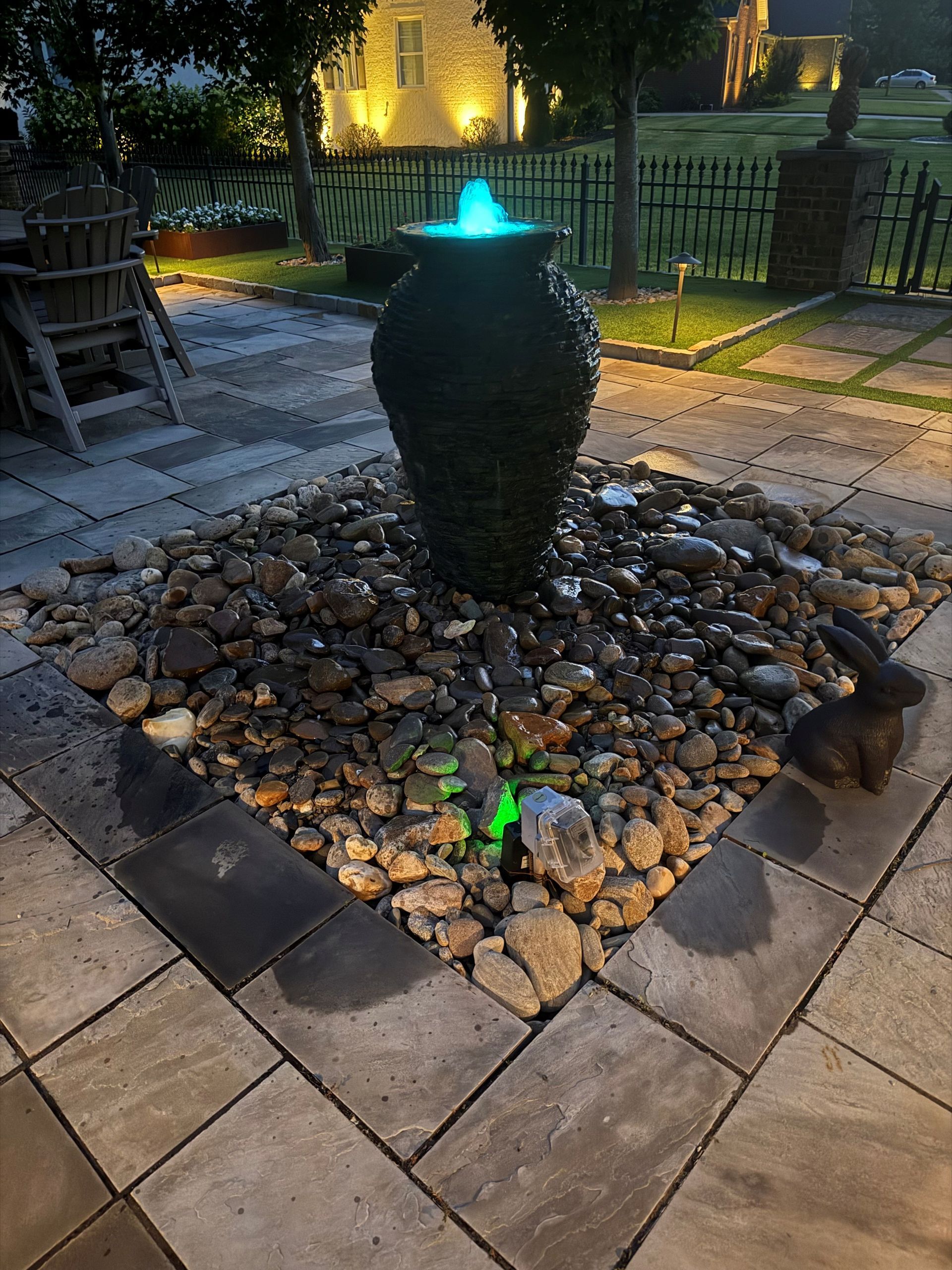Fountain with blue water, in a stone-lined bed, surrounded by a patio and garden, lit at dusk.